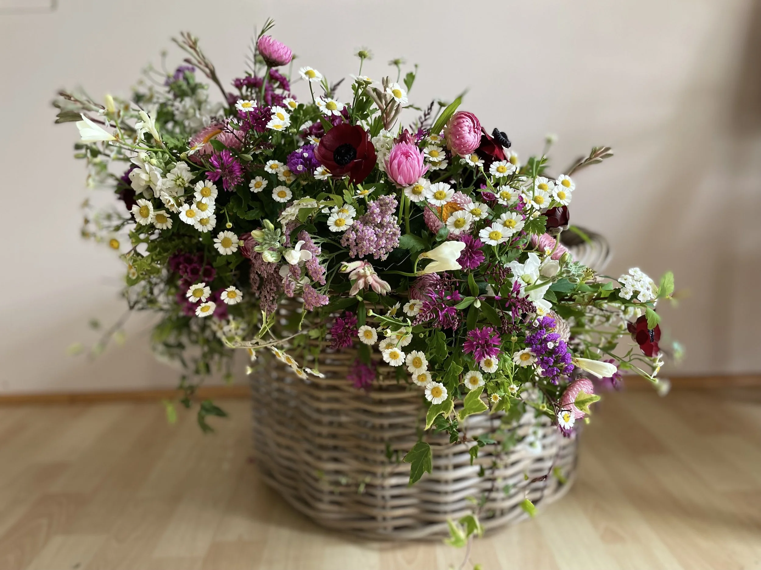 Colorful flower arrangement with daisies, poppies, primroses, and other flowers in a woven basket on a wooden surface.