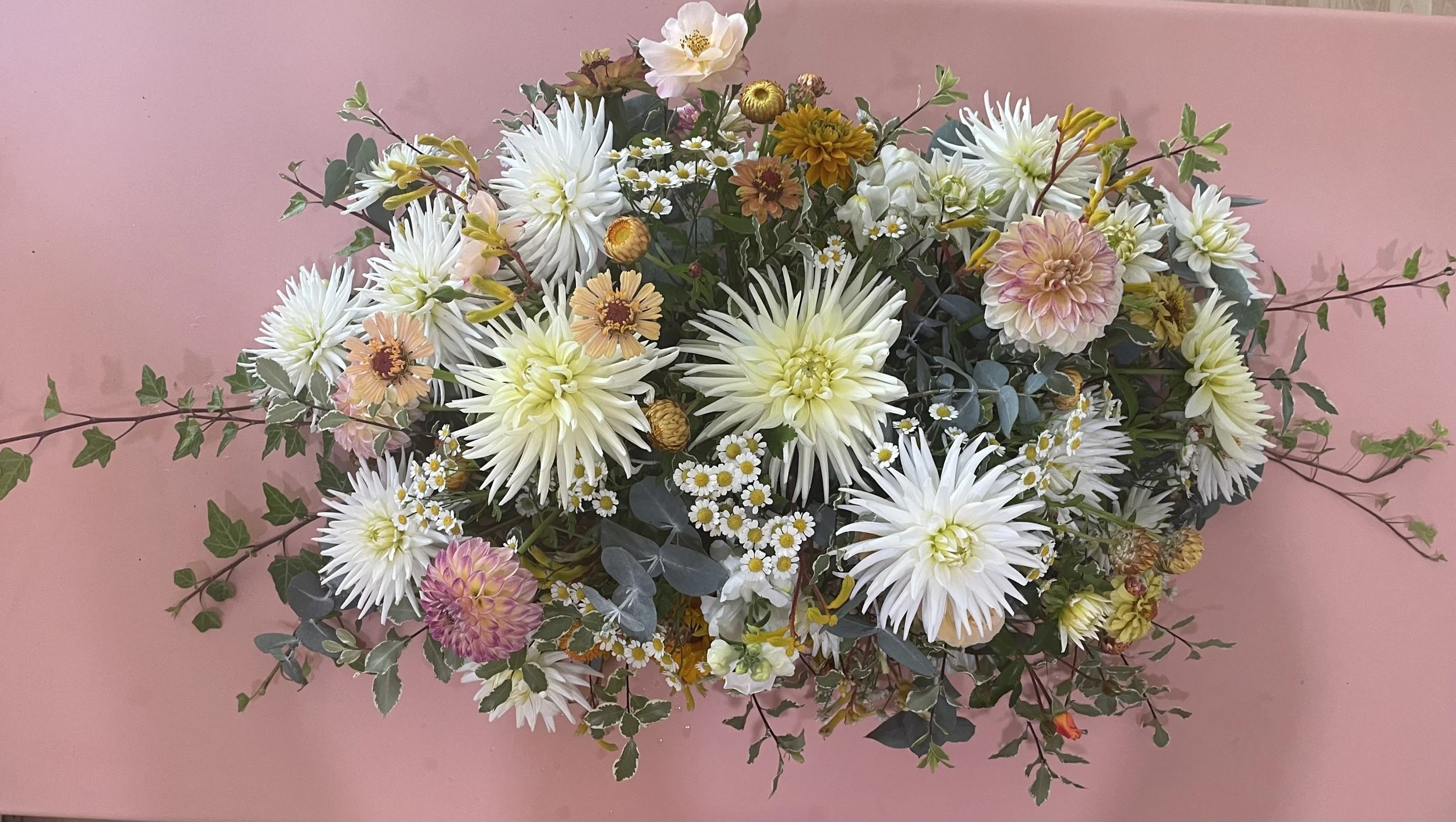A bouquet of various flowers including white dahlias, peach daisies, pink and yellow dahlias, small white daisies, and green foliage on a pink background.