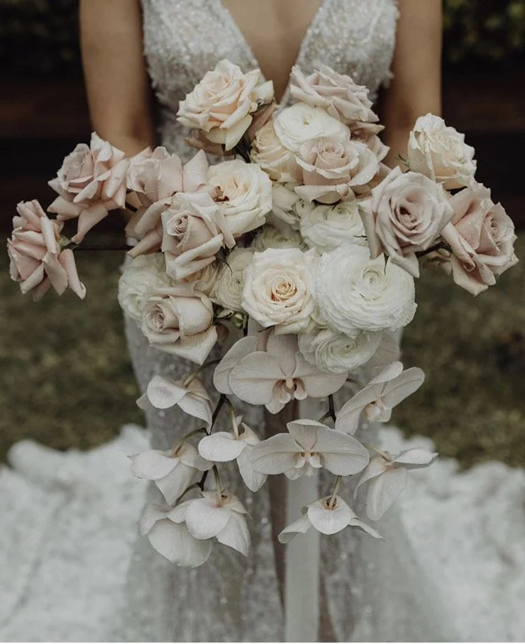 A bride holding a cascading bouquet of blush pink, cream roses, and white orchids, in front of her wedding dress.