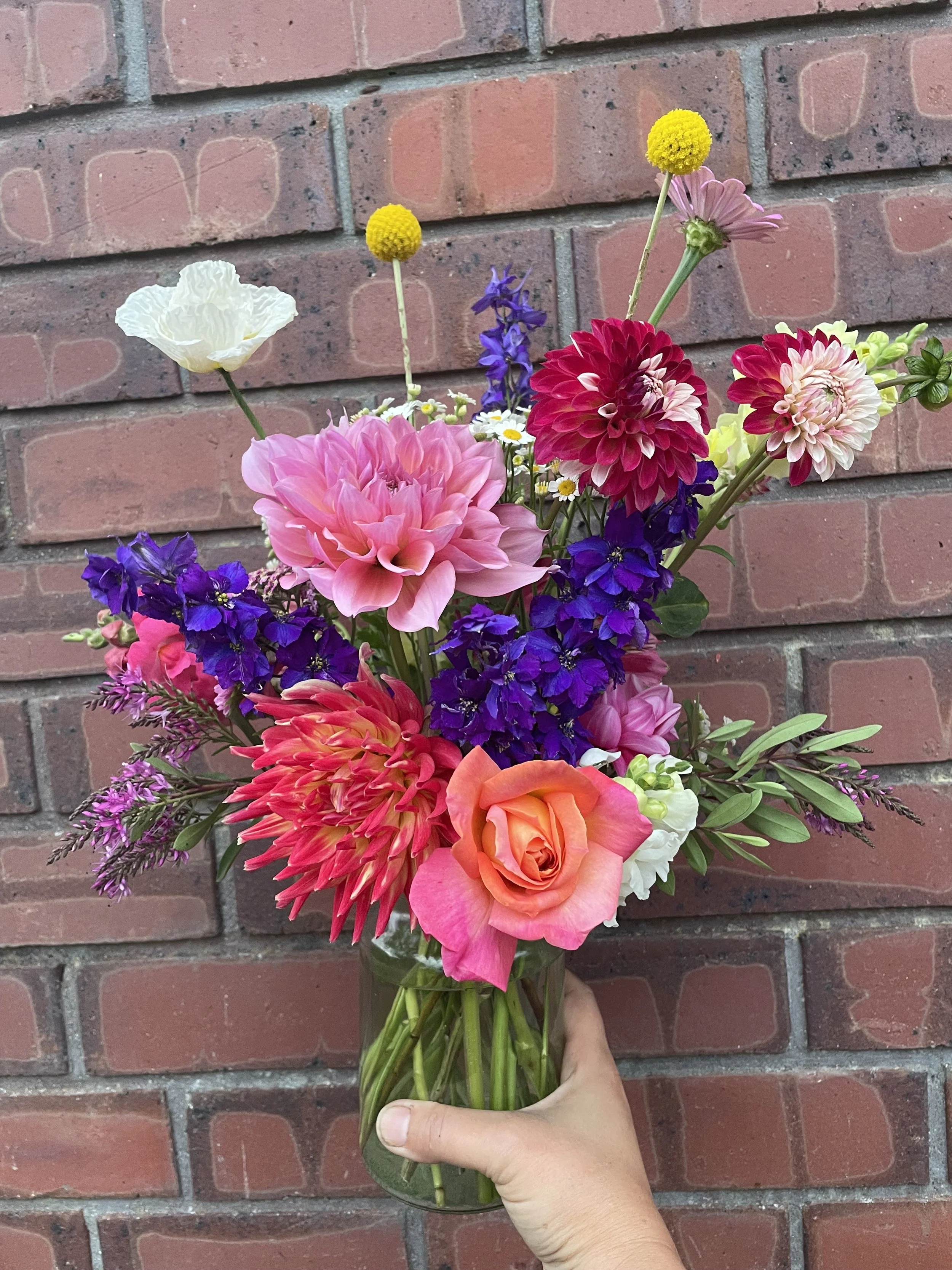 Colorful bouquet of various flowers including pink dahlias, orange rose, purple delphiniums, white poppy, yellow billy buttons, and other flowers in a glass vase held by a hand in front of a brick wall.