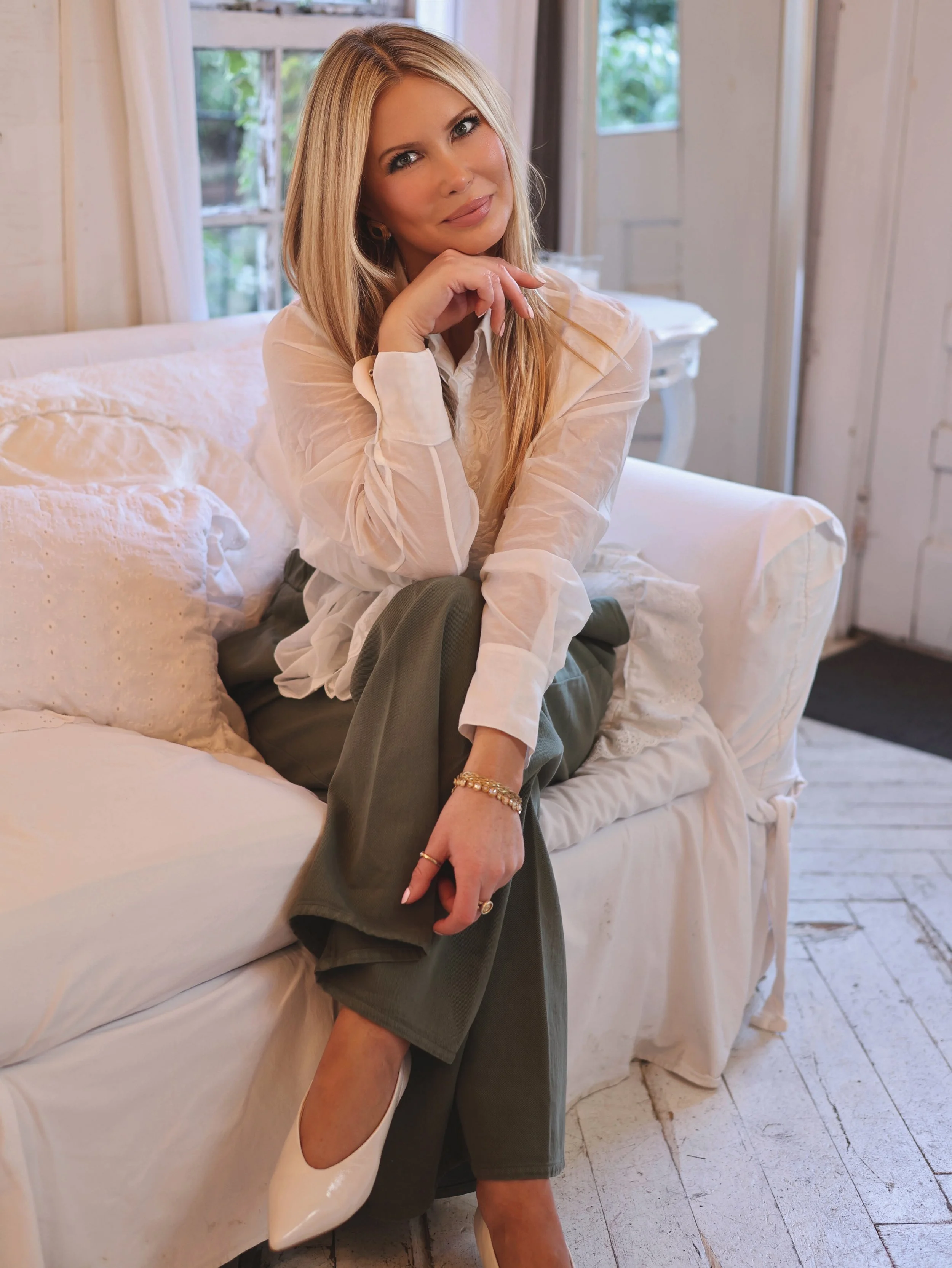 A woman with long blonde hair, wearing a sheer white blouse, olive green pants, and white high heels, is sitting on a white sofa in a cozy room with white wooden walls and windows, resting her chin on her hand and smiling softly.