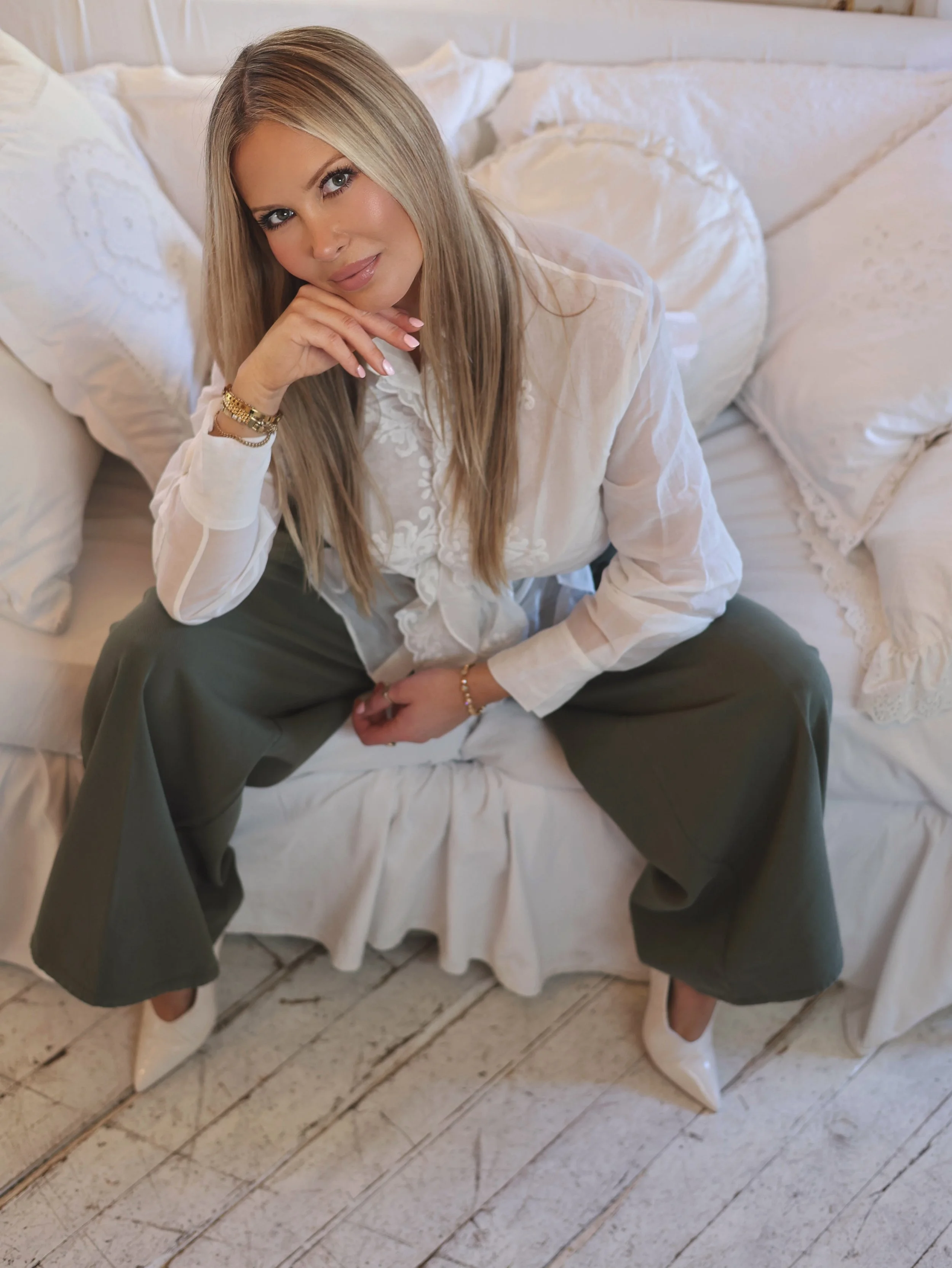 A woman with long blonde hair sitting on a white sofa with white pillows, wearing a white blouse, green wide-leg pants, and white heels, looking at the camera with a slight smile, resting her chin on her hand.