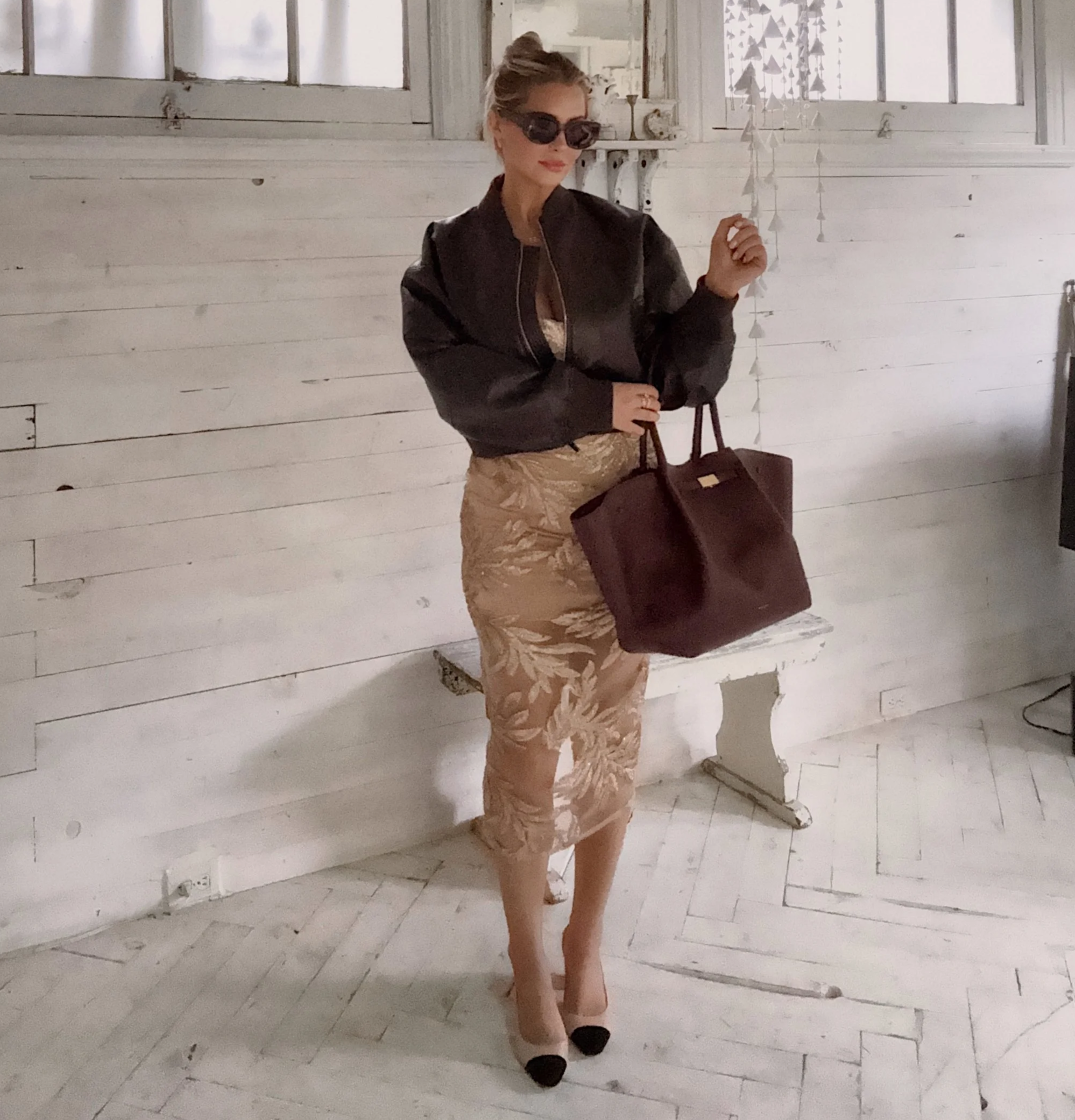 A woman standing indoors against a white wooden wall, wearing black sunglasses, a black jacket, a beige patterned dress, and black and beige high heels, holding a large brown handbag.