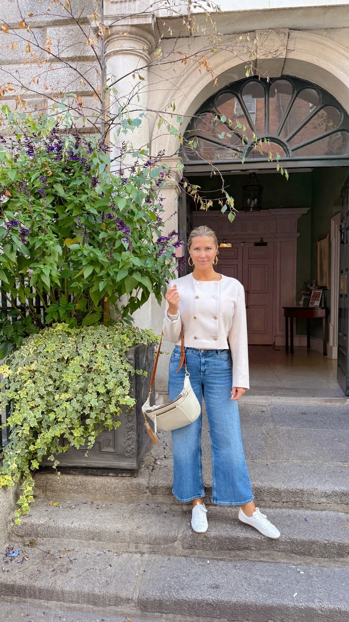 A woman standing on steps in front of a stone building entrance, with leafy plants and purple flowers on her left, wearing a white jacket, wide-legged jeans, white sneakers, and carrying a beige shoulder bag.