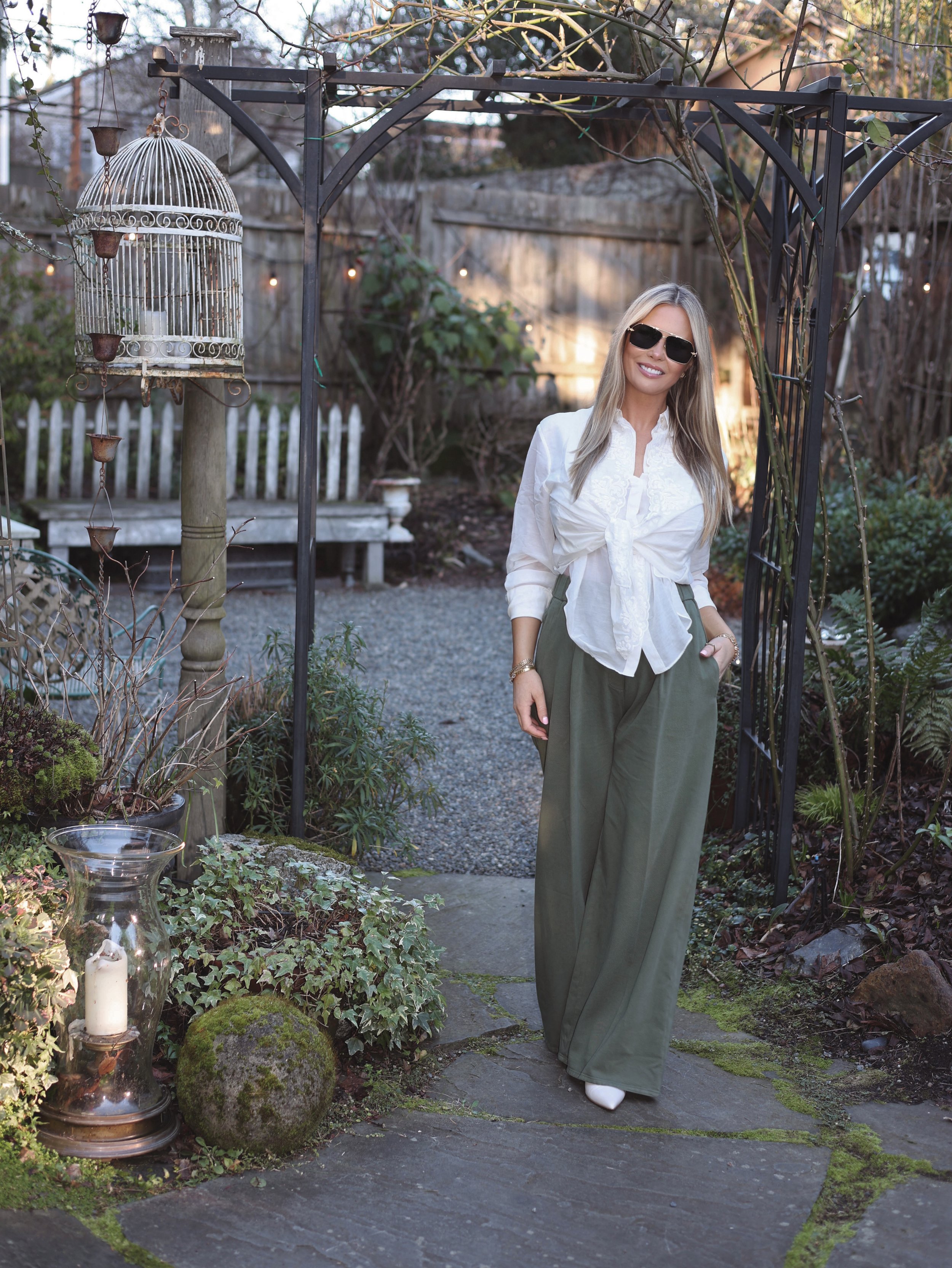 A woman with long blonde hair wearing sunglasses, a white blouse, and green wide-leg pants standing in a garden under a black metal archway.