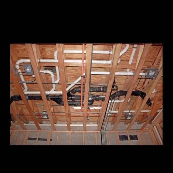 View of a building's unfinished ceiling showing wooden framing and various electrical and plumbing pipes and ducts.