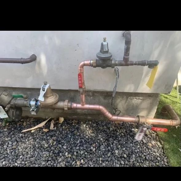 Outdoor plumbing system with copper and plastic pipes connected to a water meter and various valves, installed against a concrete wall on gravel ground.
