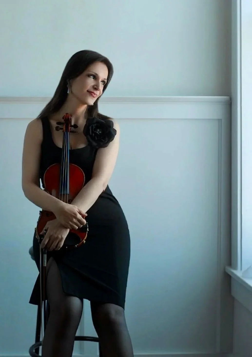 A woman, Marija Bubanji, in a black dress holding a violin indoors near a window, with a contemplative expression.
