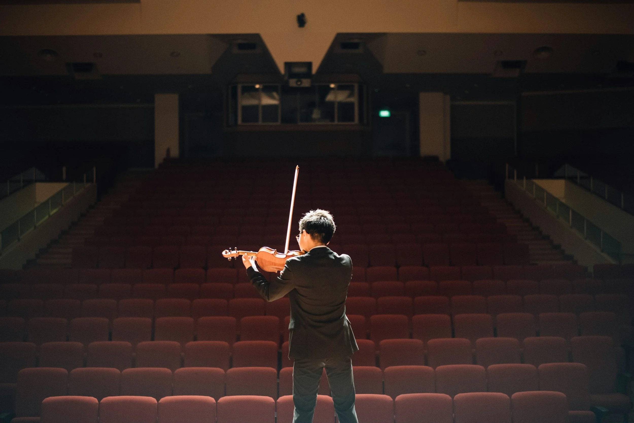 A person in a suit playing the violin on stage in an empty theater with red seats.