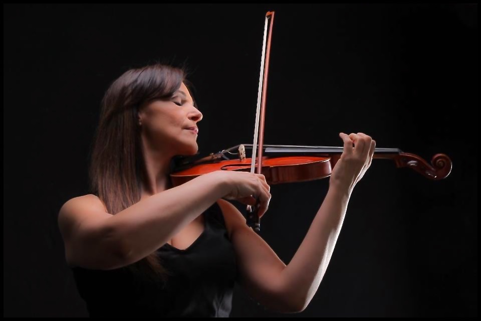 A woman, Marija Bubanji, with long brown hair playing a violin, wearing a black sleeveless top, against a black background.