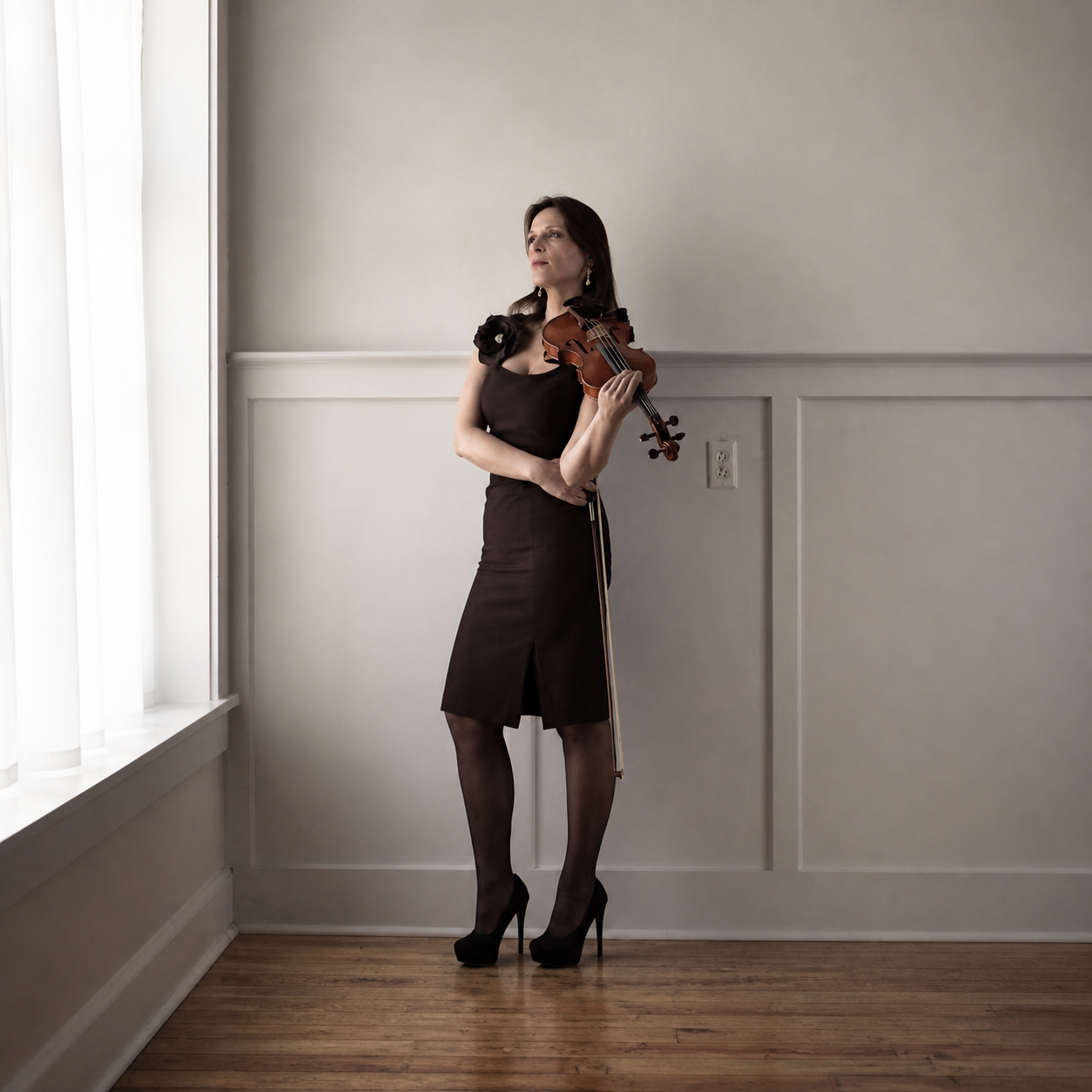 A woman, Marija Bubanji,  in a black dress and high heels standing with a violin in a room with white walls and hardwood floor.