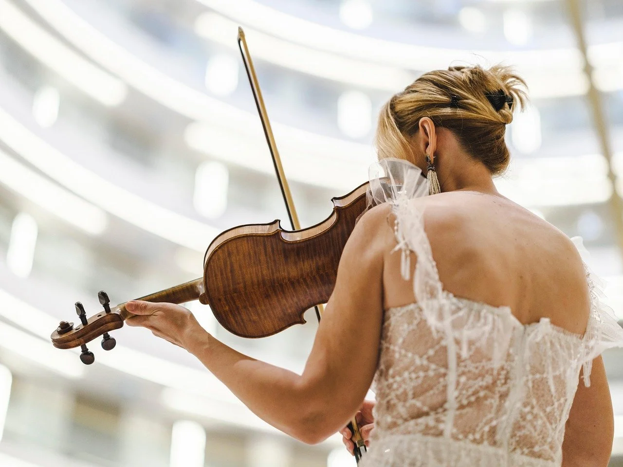 A woman with blonde hair styled in an updo, wearing a lace dress and earrings, is playing a violin in a bright, modern indoor setting with curved balconies.