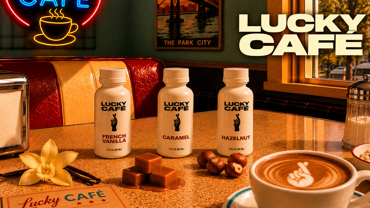 Interior of Lucky Cafe featuring three bottles of flavoring syrups labeled French Vanilla, Caramel, and Hazelnut, a latte with latte art, caramel squares, hazelnuts, a flower, and a neon sign with a coffee cup.