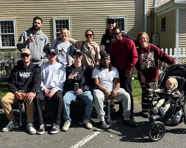 Group of ten people, adults and teenagers, posing outdoors on a sidewalk next to a yellow house with white trim. One person is pushing a child in a stroller. They are smiling and holding drinks.