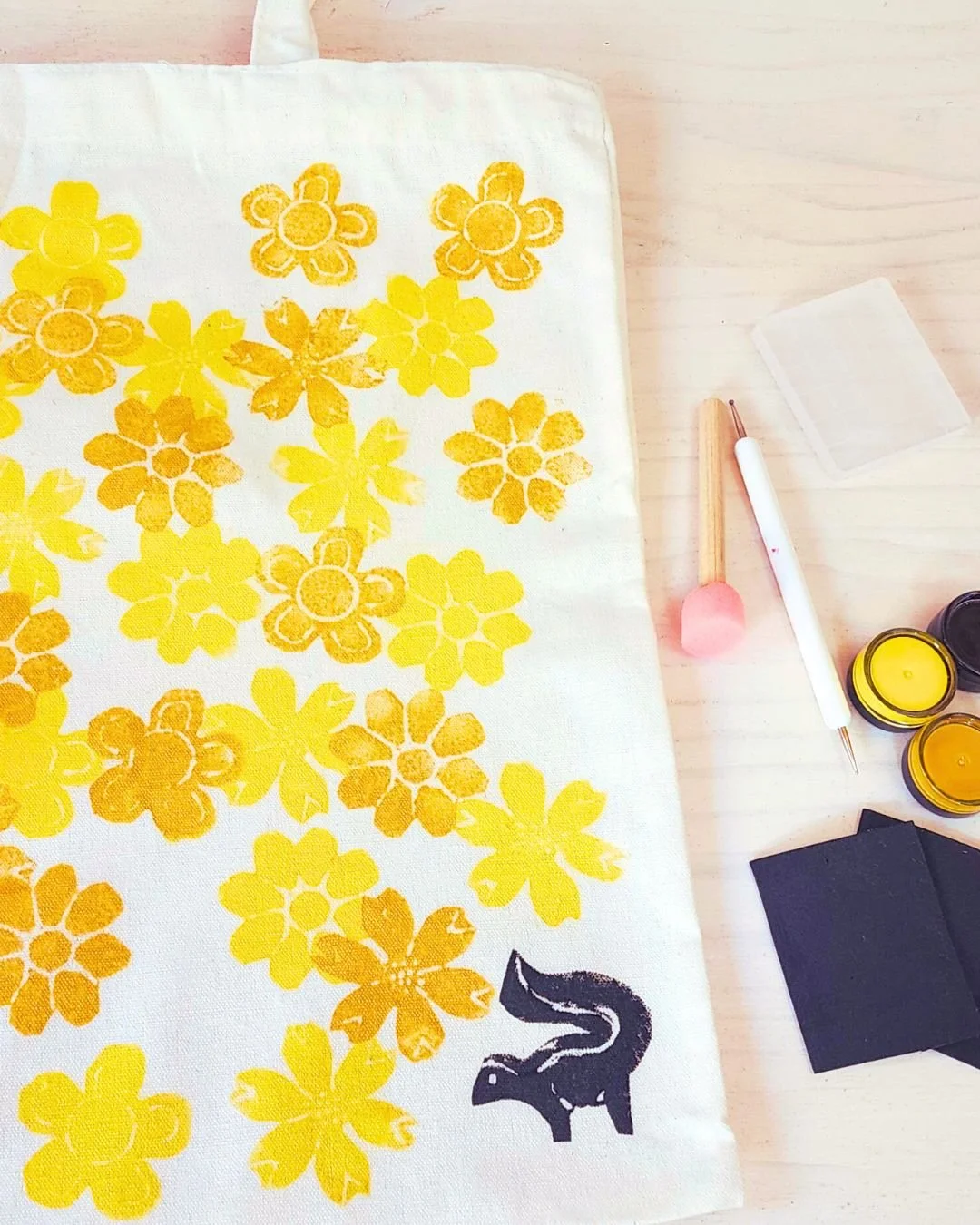 Fabric with yellow flower patterns, black animal silhouette on bottom right, and craft tools including paint jars, brush, pen, tissue, and black fabric on light wooden surface.