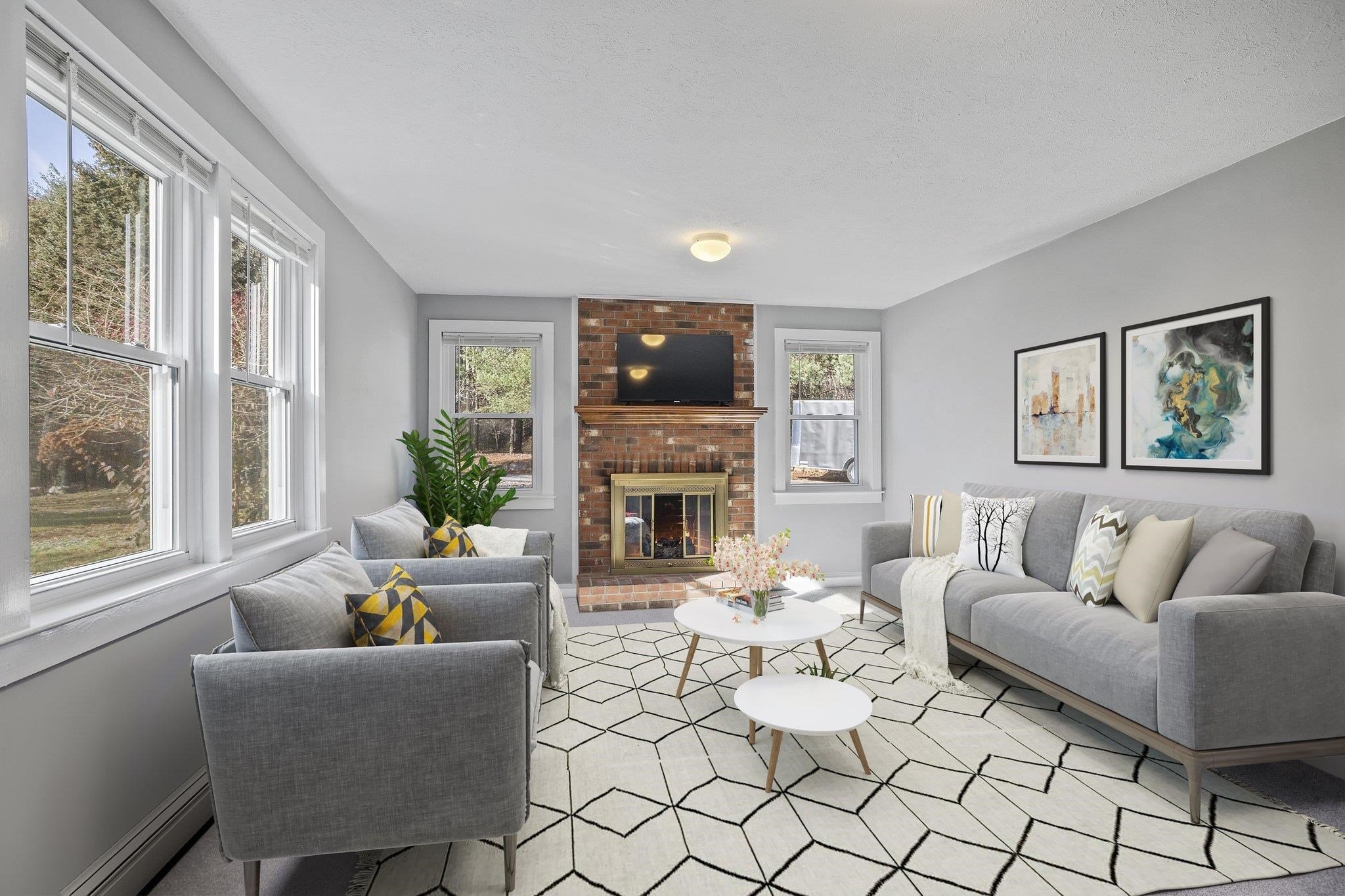 Modern living room with light gray walls, two gray armchairs, a gray sofa with decorative pillows, a geometric patterned rug, a white coffee table, and a brick fireplace. A TV is mounted above the fireplace, and two framed artworks hang on the wall.