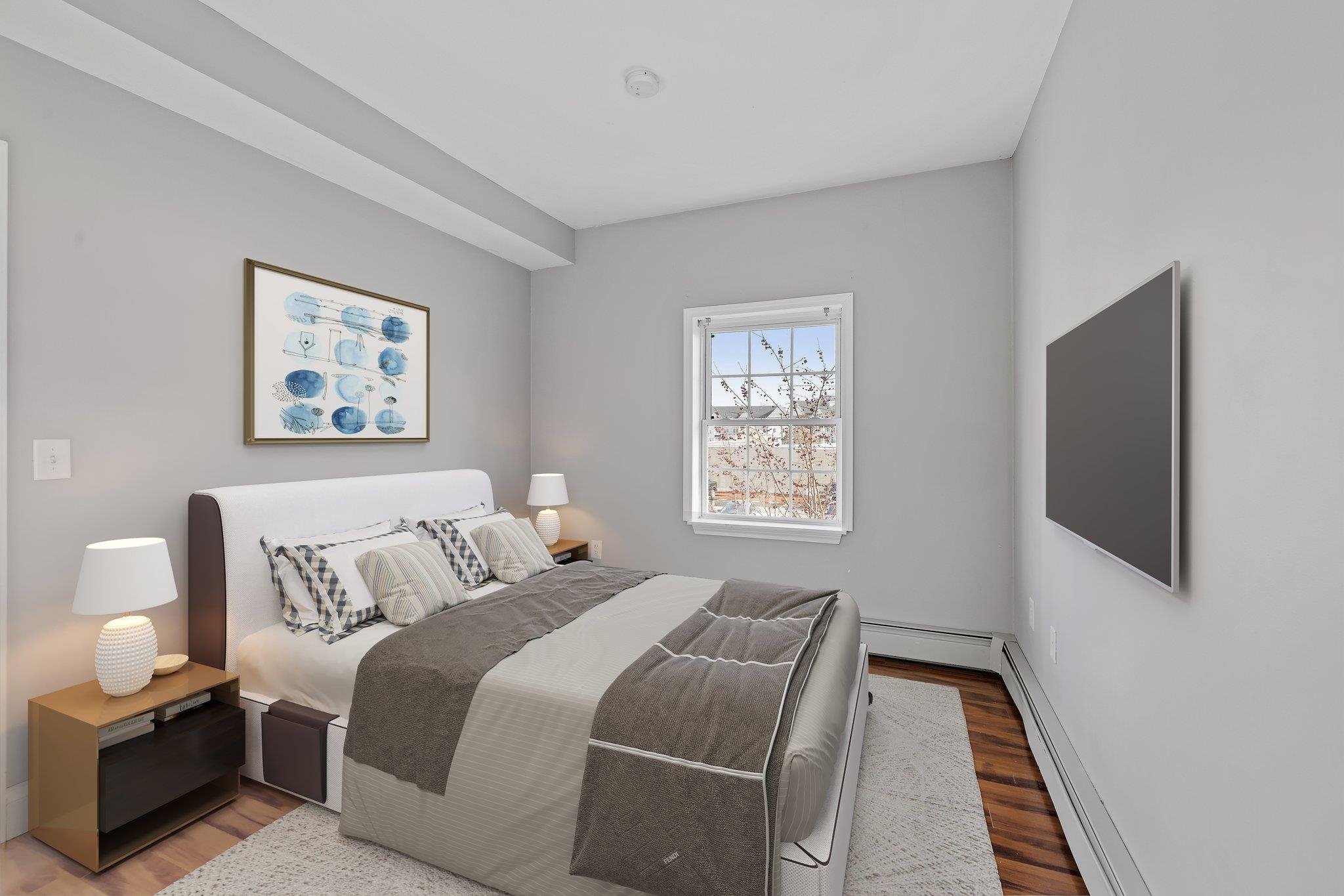 Modern bedroom with gray walls, featuring a double bed adorned with striped pillows and a neutral comforter, flanked by two bedside lamps on nightstands. A large abstract painting hangs above the bed, and a window with a view of trees is on the side.