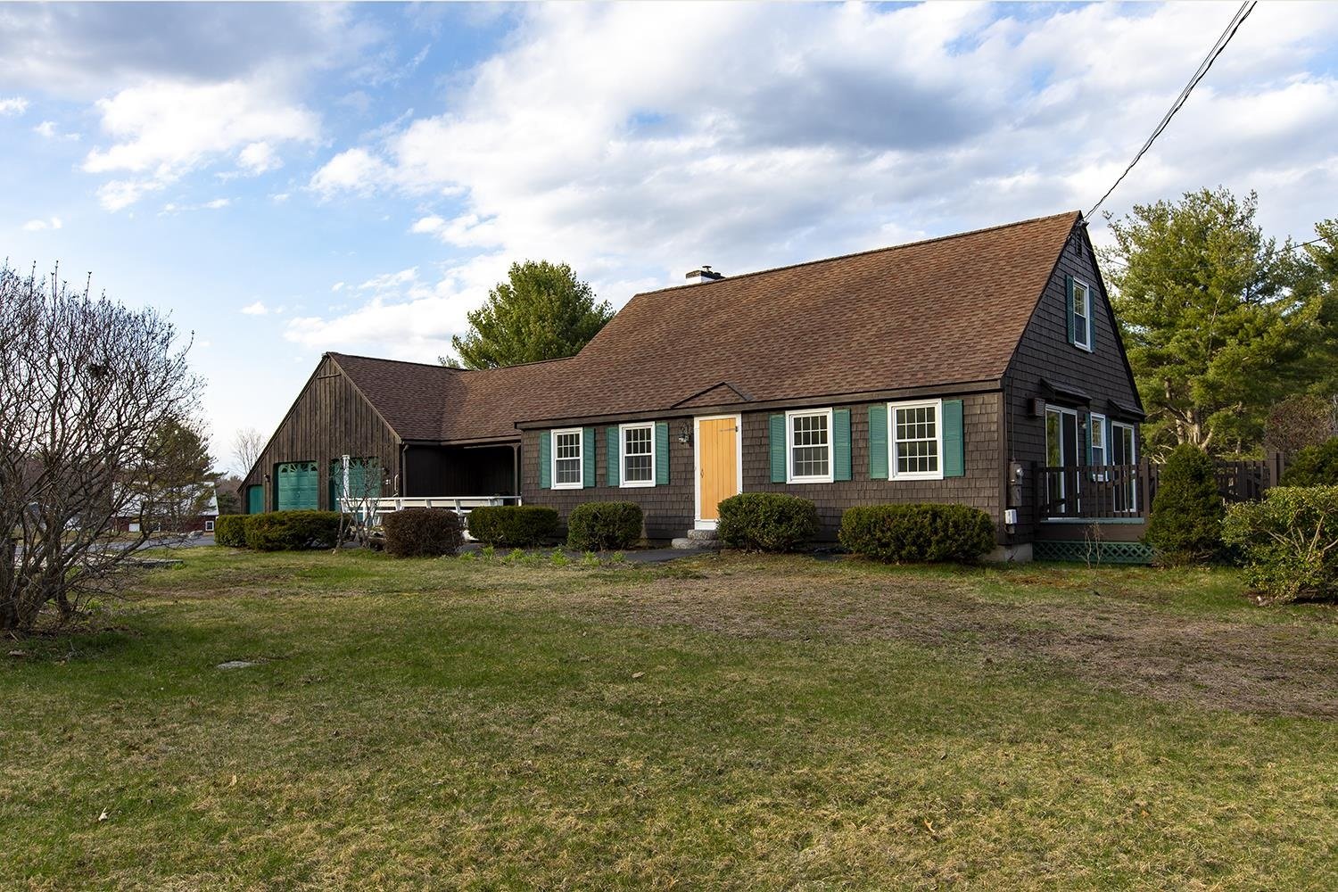 A suburban house with brown shingles, a sloped roof, green window shutters, and a well-maintained lawn under a partly cloudy sky.