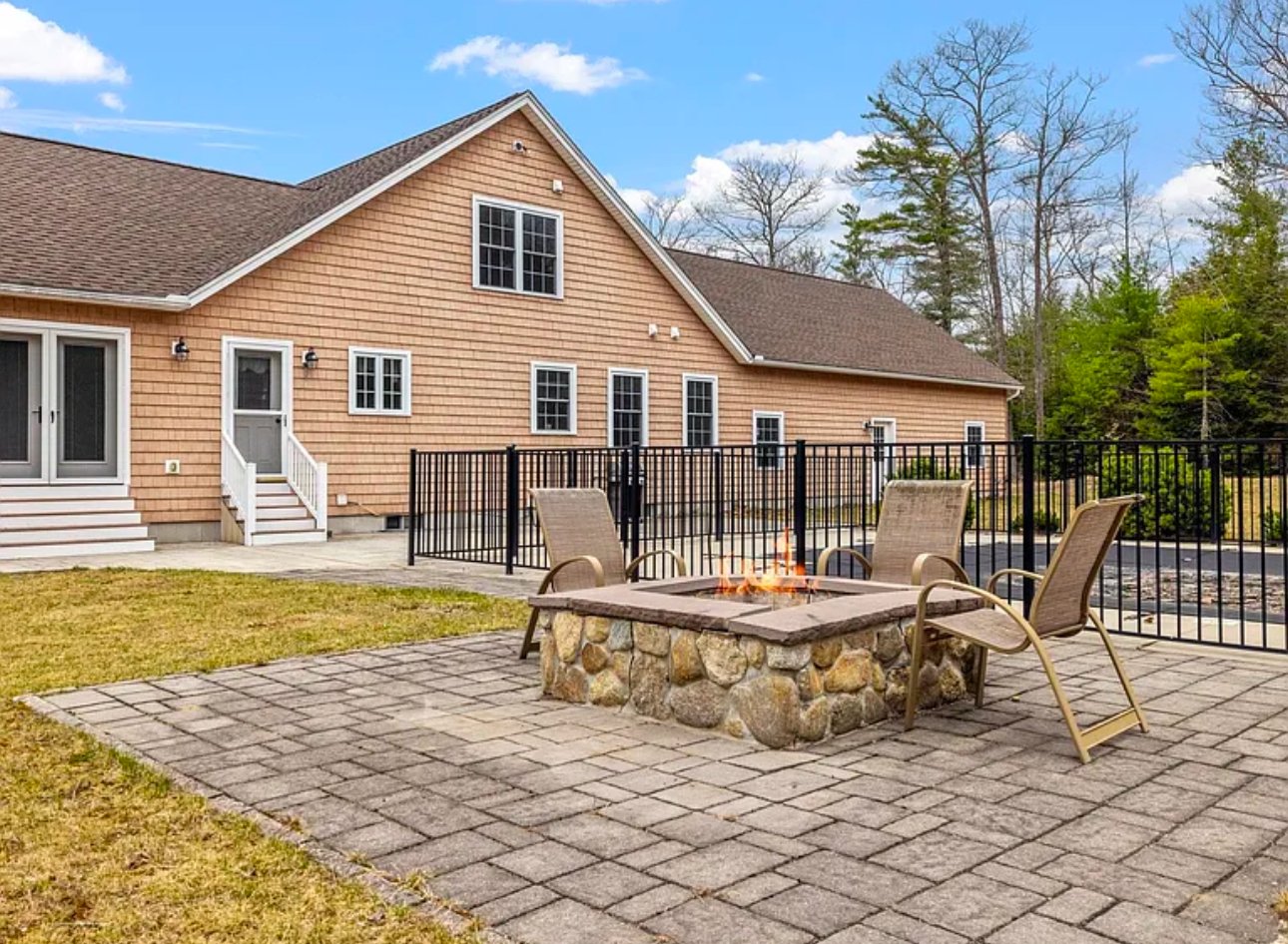 A backyard with a large tan house, stone fire pit, and patio chairs on a paved area.