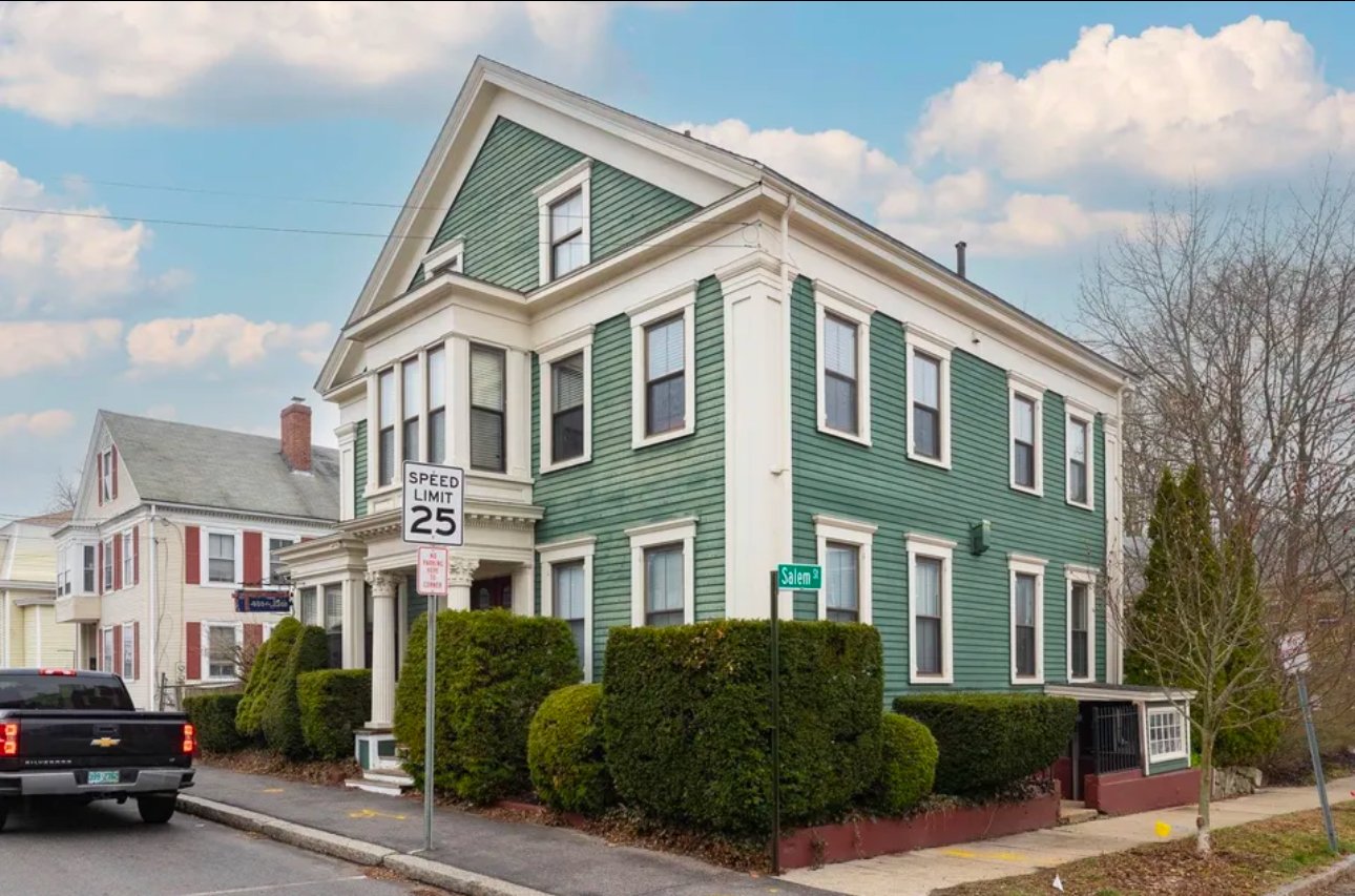 Green two-story house with bay windows and a speed limit sign in a suburban neighborhood.