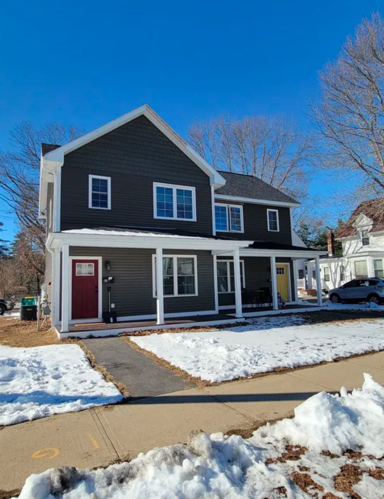 Modern two-story house with dark siding, red and yellow doors, covered porch, surrounded by snow and a clear sky.