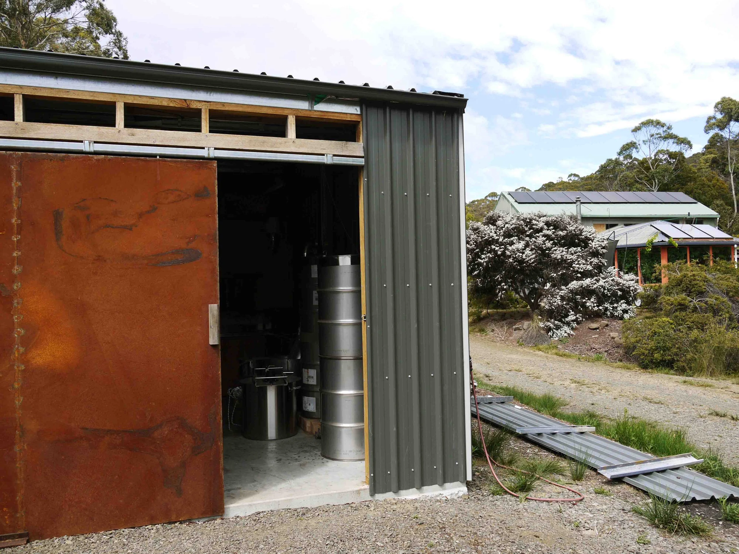 Partially constructed shed with a rust-colored sliding door and stainless steel distillation barrels inside. In the background, there is a house with solar panels, surrounded by trees and flowering bushes.