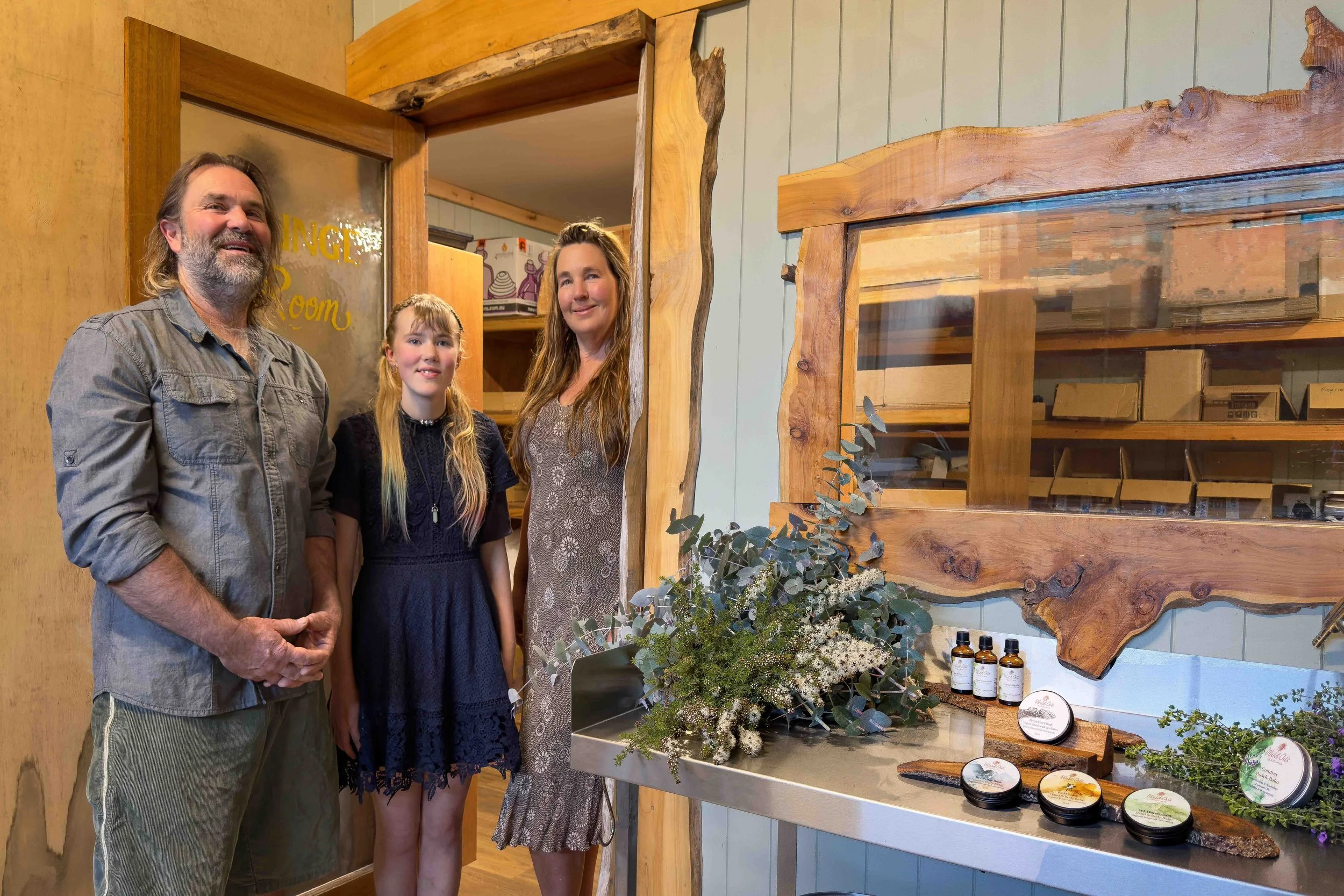 A family of three standing in a wooden doorway of a workshop, with stainless steel benches, plant material and tins and bottles of aromatherapy products.