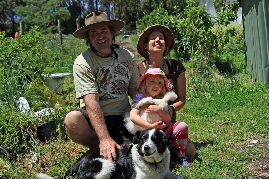 A man patting a black and white dog, a woman and young child holding a white chicken, in a vegetable garden.