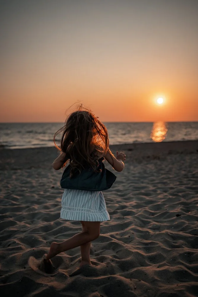 Ein Mädchen läuft am Strand bei Sonnenuntergang, das Meer im Hintergrund, die Sonne spiegelt sich im Wasser.
