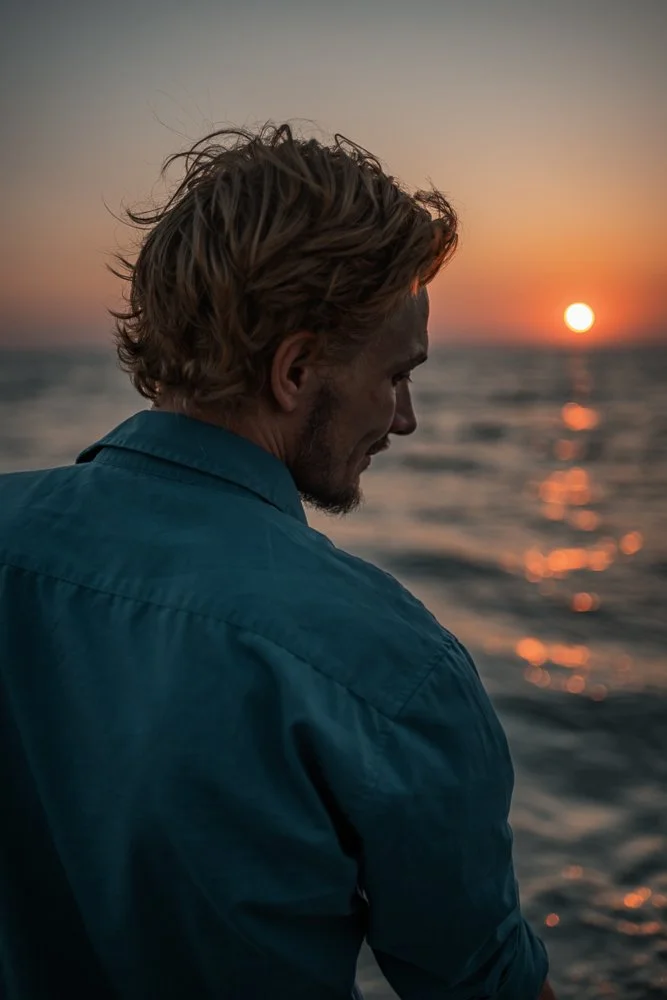 Ein Mann mit lockigem, blondem Haar schaut nachdenklich auf das Meer, während die Sonne im Horizont untergeht.
