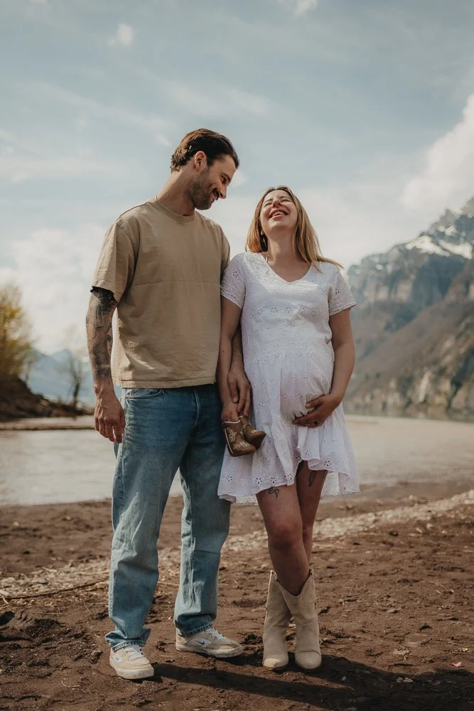 Ein Paar steht an einem Flussufer in einer bergigen Landschaft, die Frau ist schwanger und trägt ein weißes Kleid, der Mann trägt ein beige T-Shirt und Jeans.