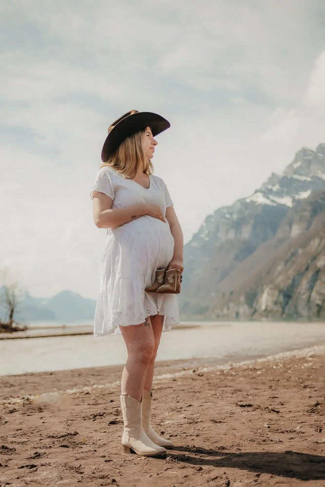 Eine schwangere Frau in einem weißen Kleid, einem schwarzen Cowboyhut und braunen Cowboy-Stiefeln, die in einer Landschaft mit Bergen im Hintergrund steht, hält ein Paar braune Stiefel in der Hand.