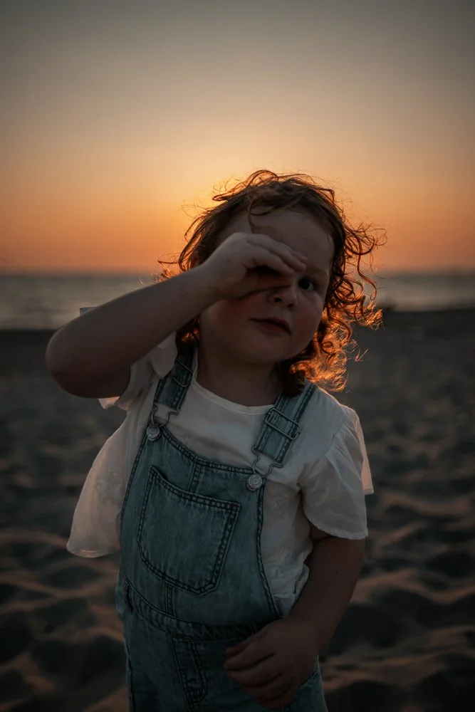Kleines Mädchen mit lockigem Haar steht am Strand bei Sonnenuntergang, trägt Jeanskleidung, hält eine Hand vor ein Auge.