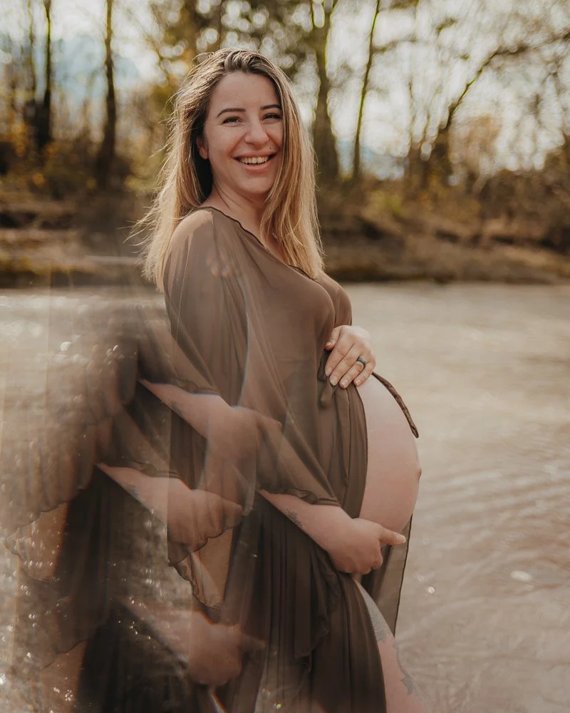 Eine schwangere Frau mit langem Haar, die im Fluss steht und sich über ihr Babybauch freut, während sie draußen in der Natur steht.