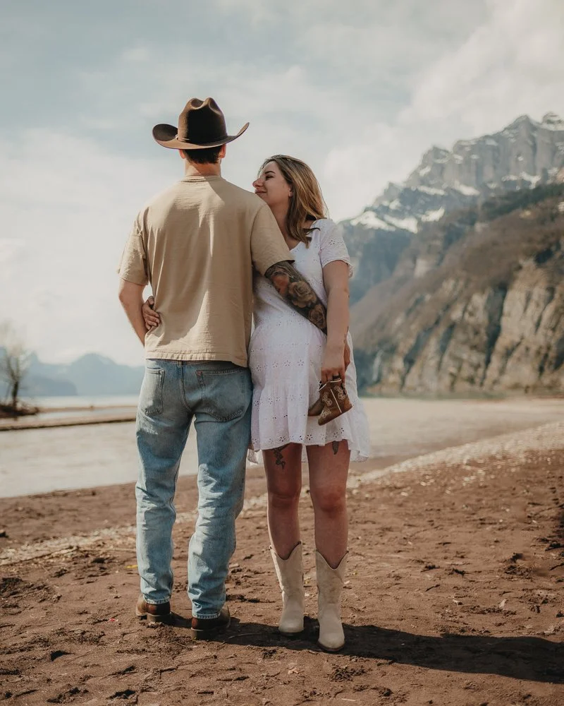 Ein Paar steht sich auf einer Strandpromenade mit Bergen im Hintergrund gegenüber, die Frau trägt ein weißes Kleid und Cowboy-Stiefel, der Mann trägt einen Cowboy-Hut und Jeans.