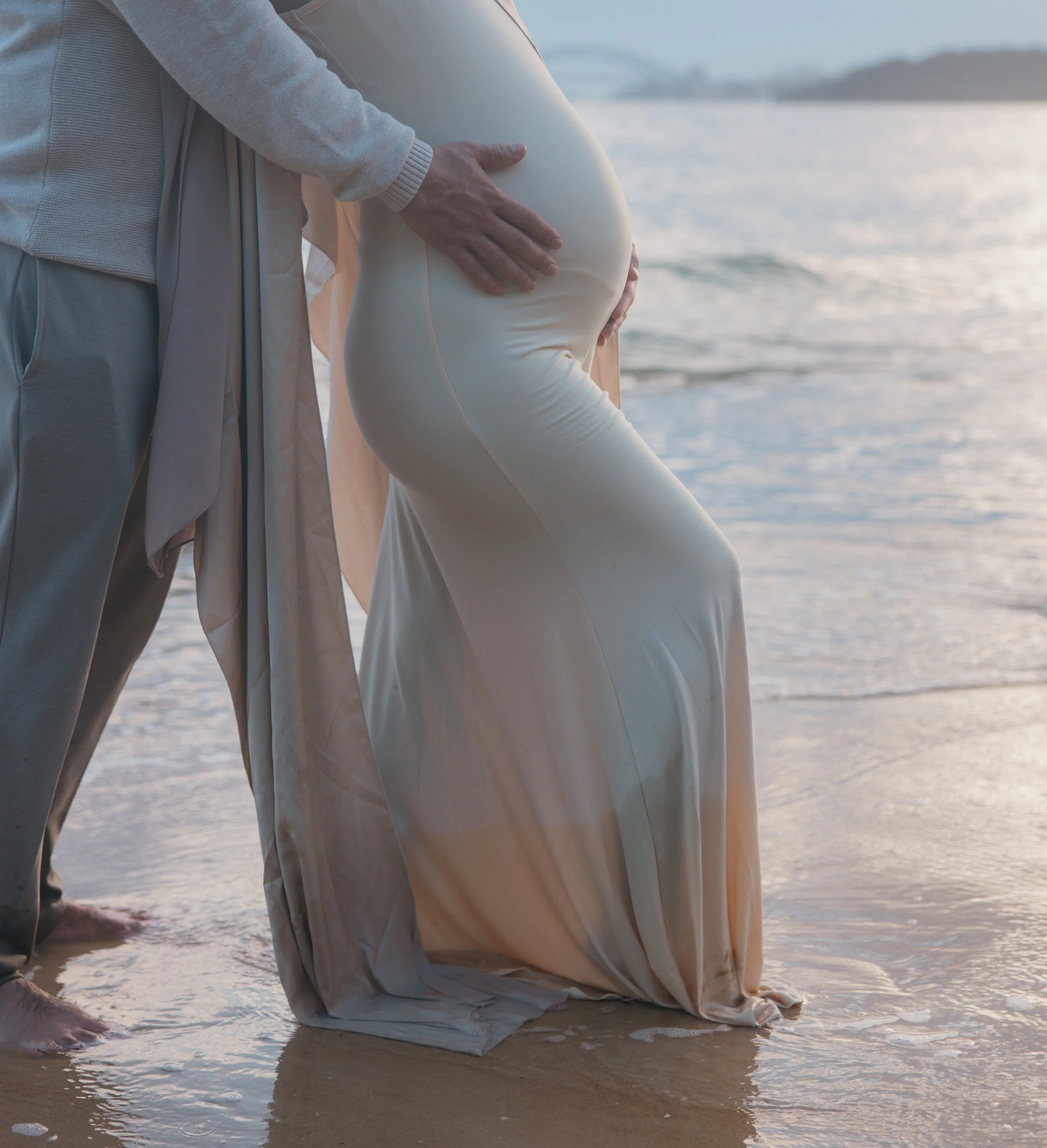 Pregnant woman in a beige dress kneeling by the ocean, holding her belly, with a man standing behind her on the beach during sunset.