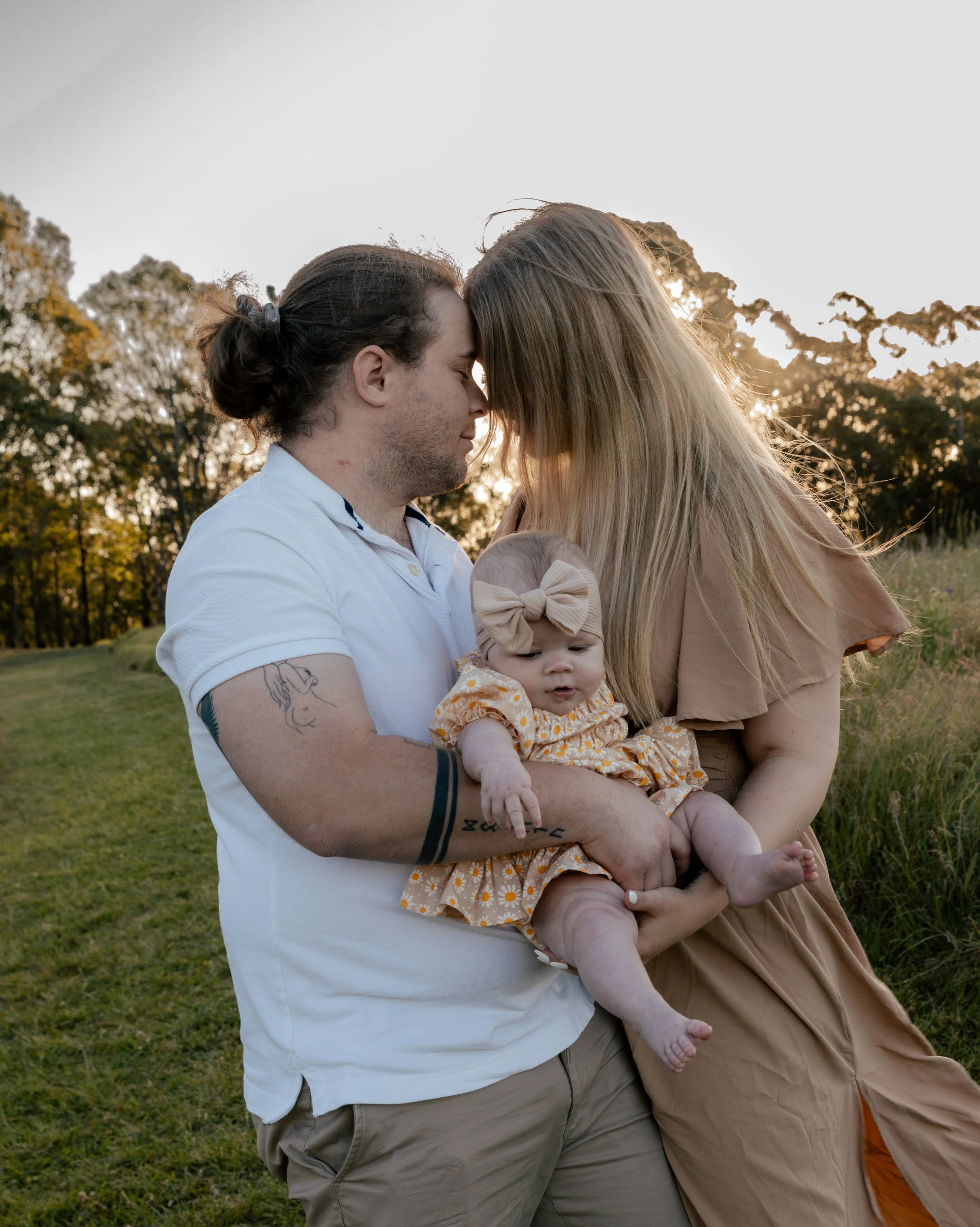 A couple holding their baby outdoors at sunset, with trees in the background.