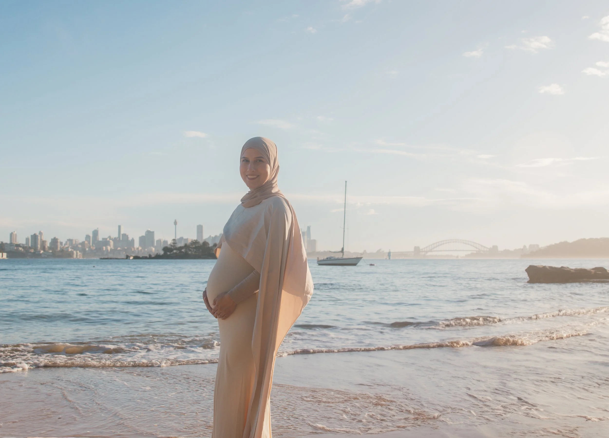 Pregnant woman wearing a beige hijab and long dress standing on a beach with the city skyline, sailboat, and bridge in the background.