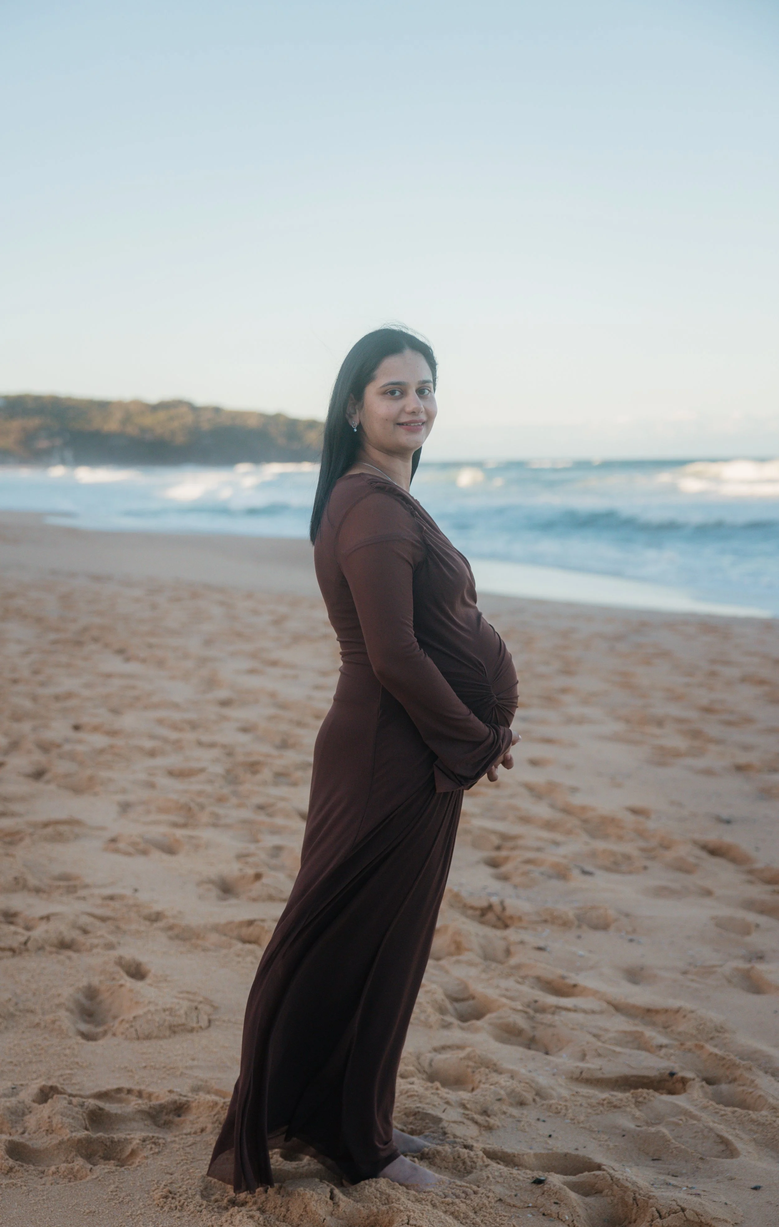A pregnant woman in a brown dress standing on a sandy beach with ocean waves and a distant shoreline in the background.