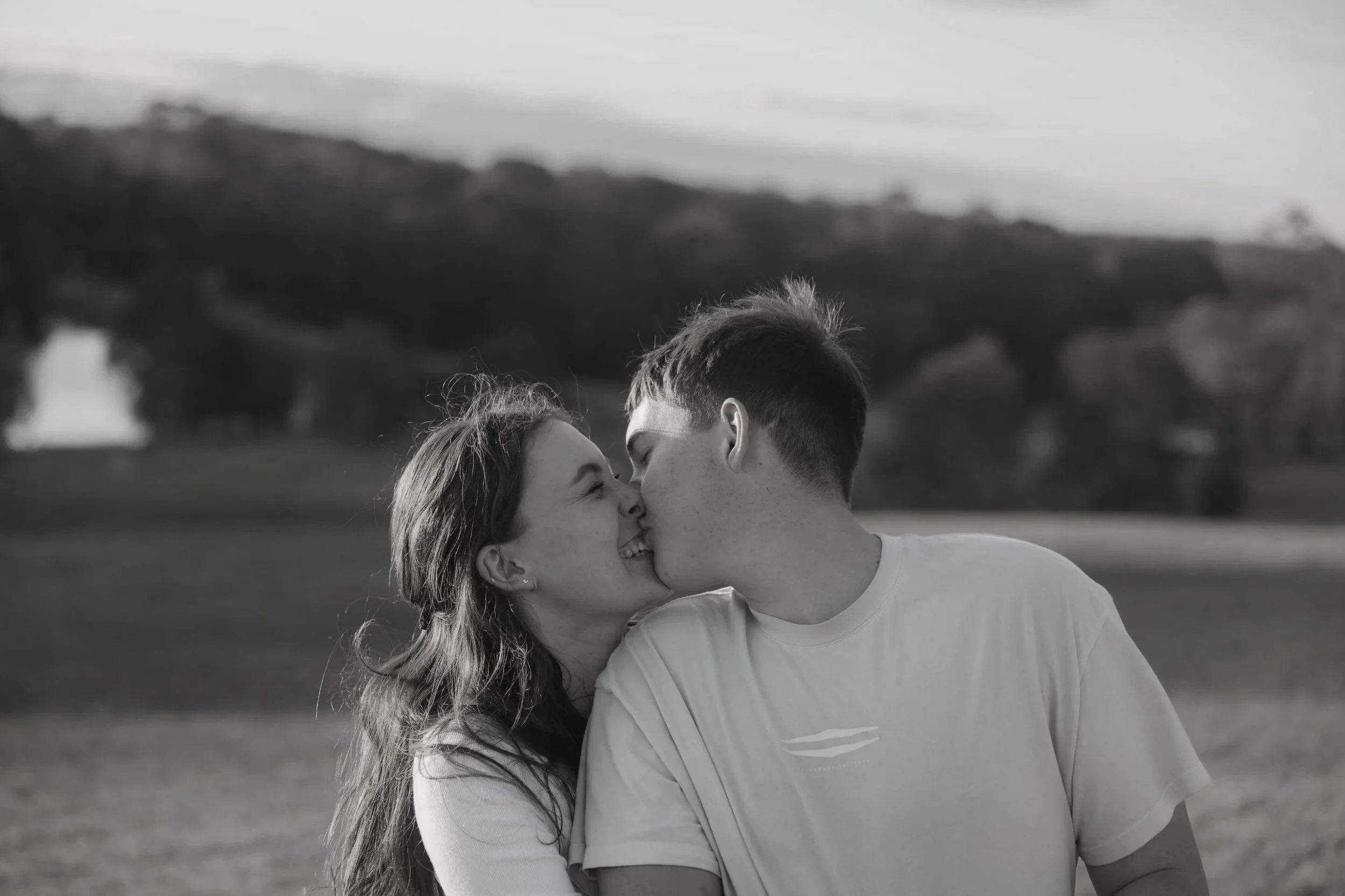 Black and white photo of a young couple kissing outdoors on a grassy field with hills and trees in the background.