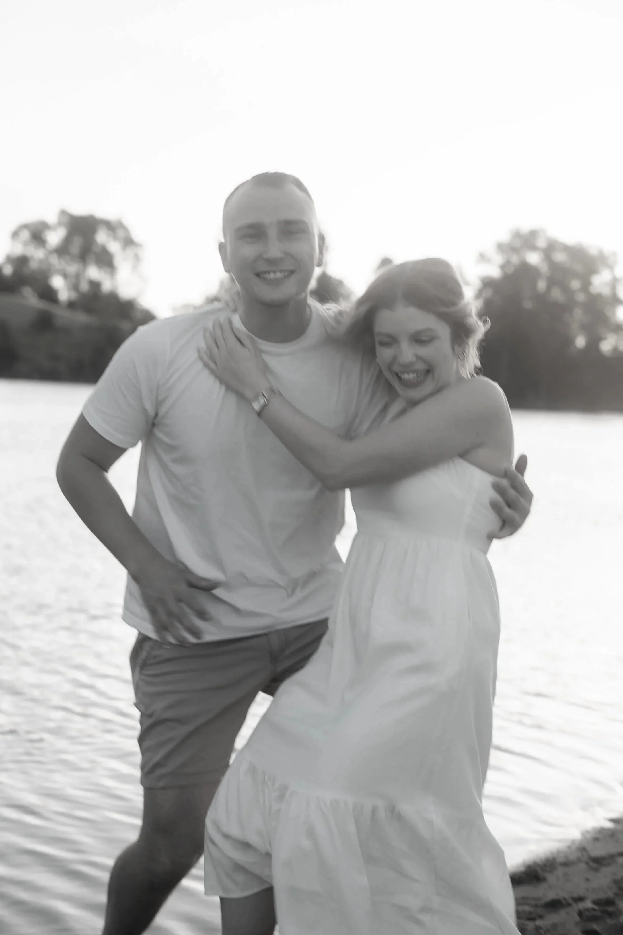 A black and white photo of a smiling young man and woman embracing by a body of water with trees in the background.