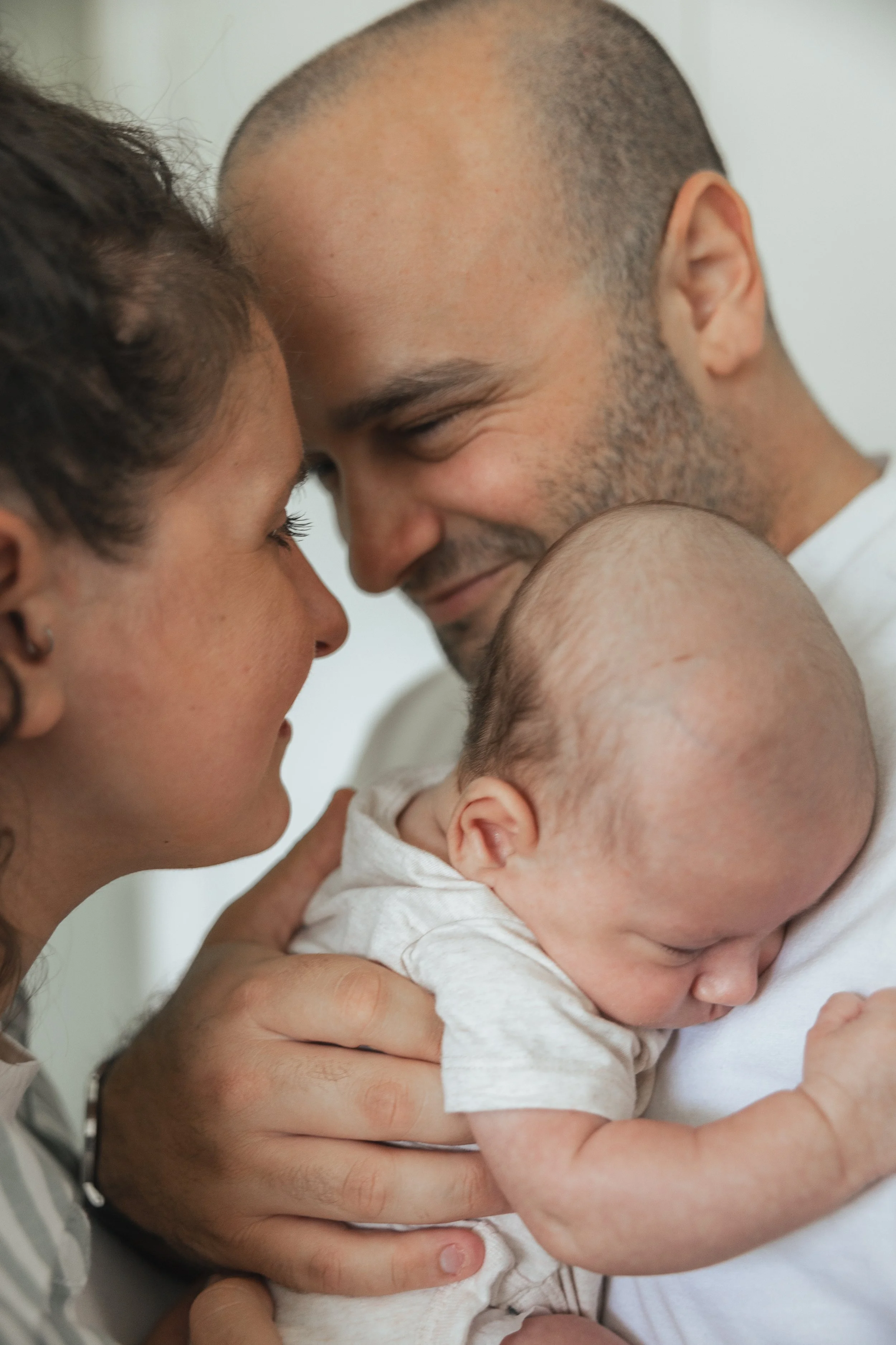 A man and woman leaning close with their foreheads touching, holding a sleeping baby with a bald head and closed eyes.