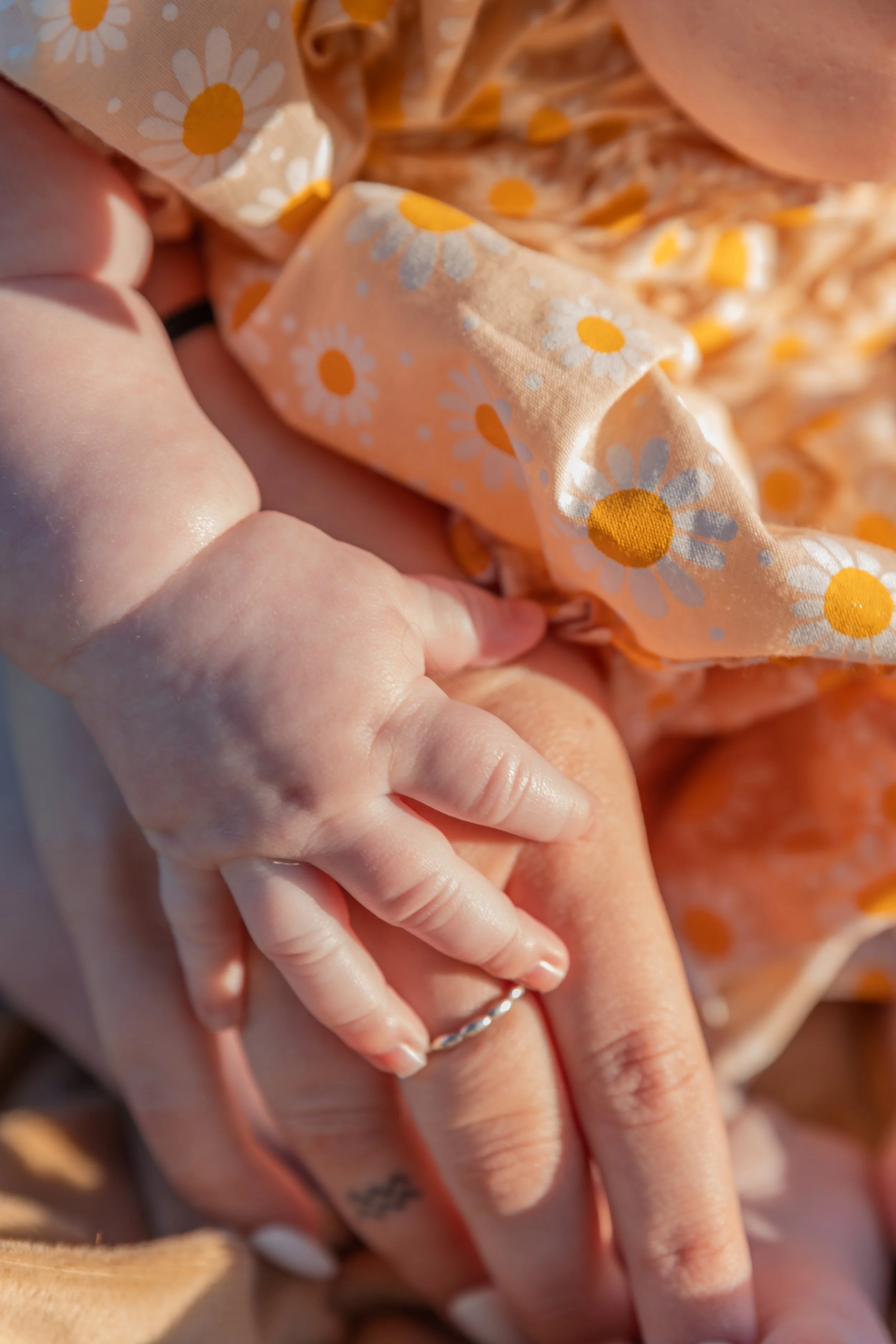 A baby's hand is holding an adult's finger, with the adult wearing a ring on their finger. The person is wearing a daisy-print, peach-colored garment.