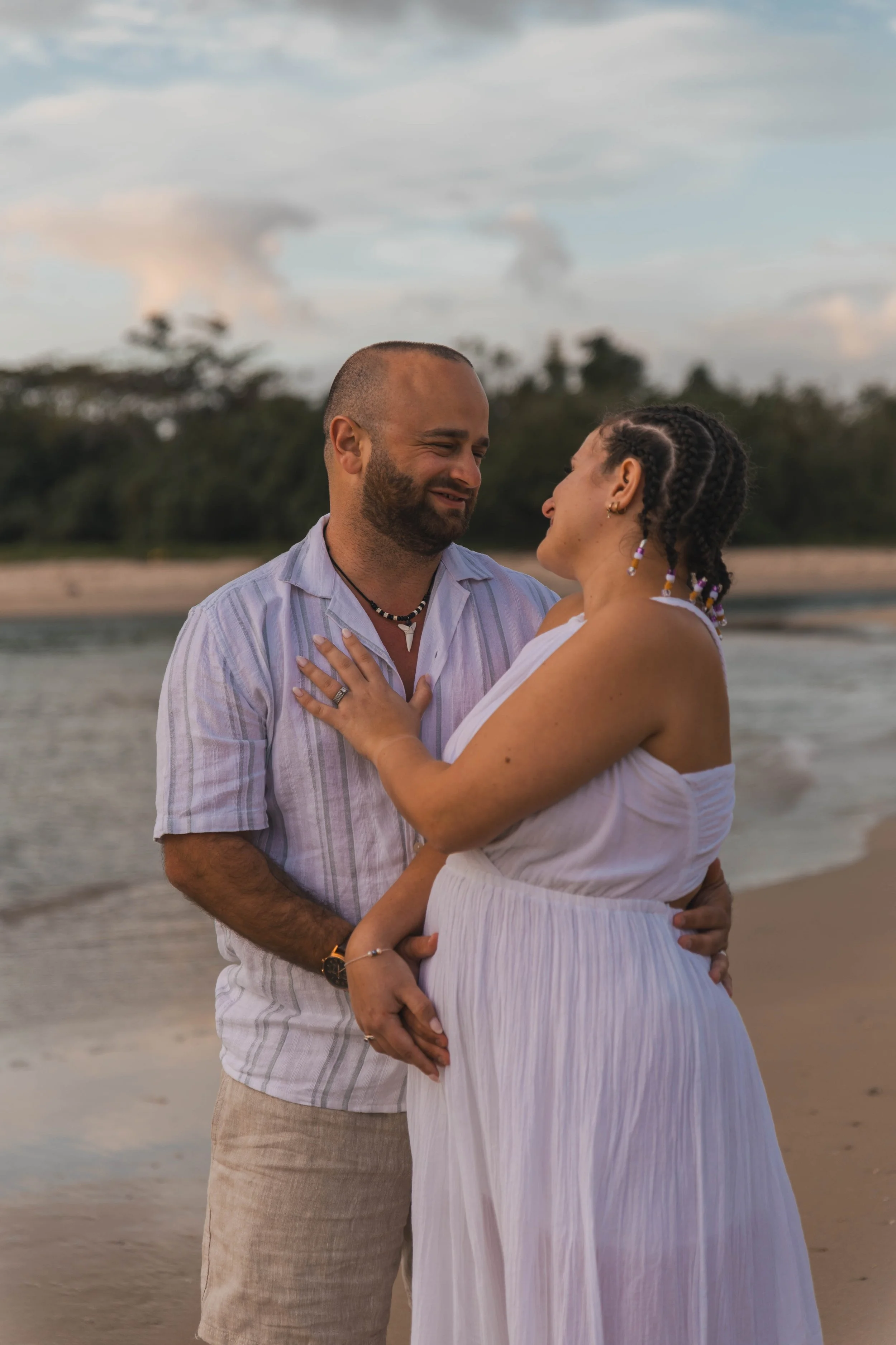 A couple embraces by the water on a beach during sunset, smiling and looking at each other lovingly.
