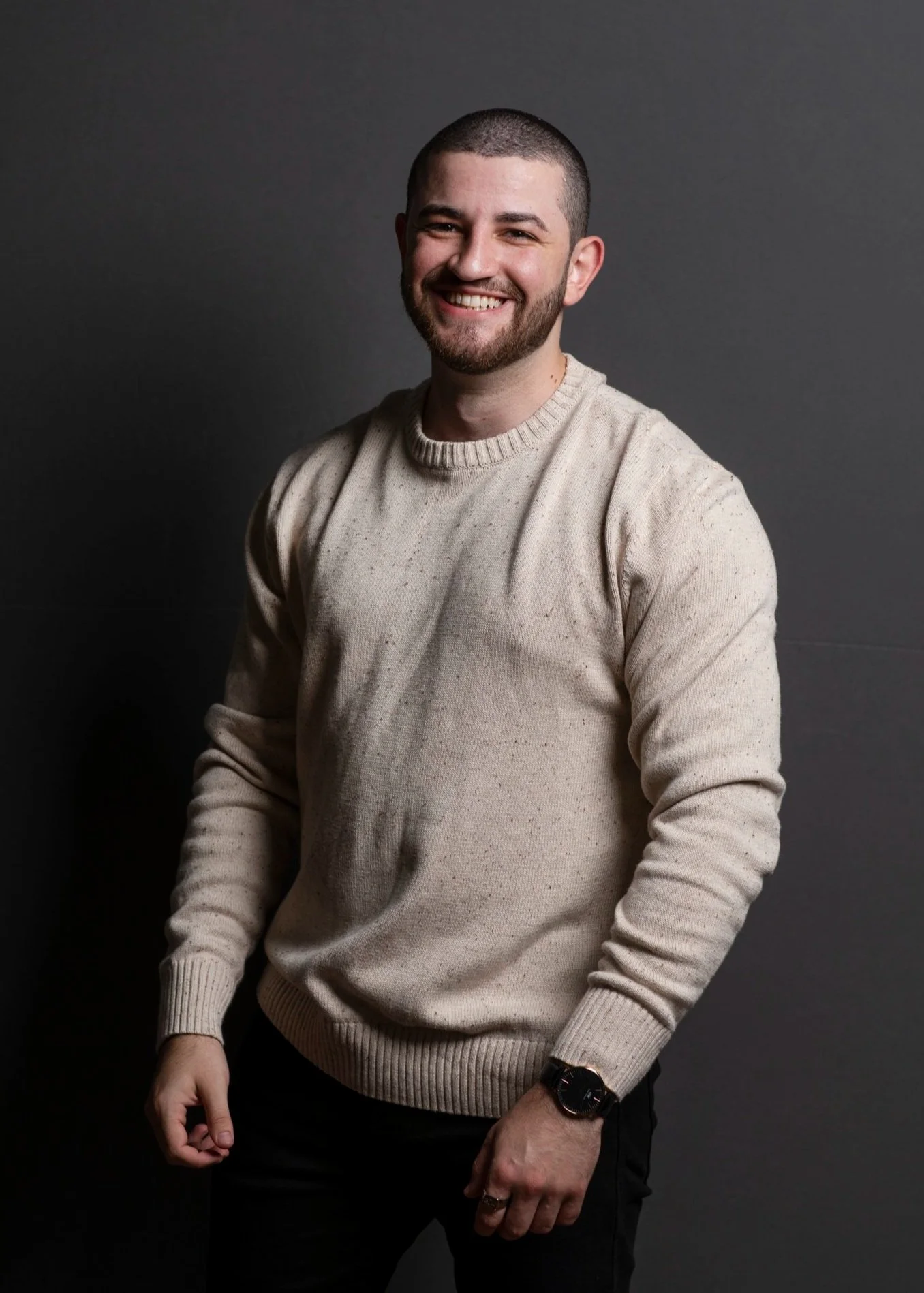 Young man with a beard and short hair smiling, wearing a beige sweater and black pants, standing against a dark gray background.