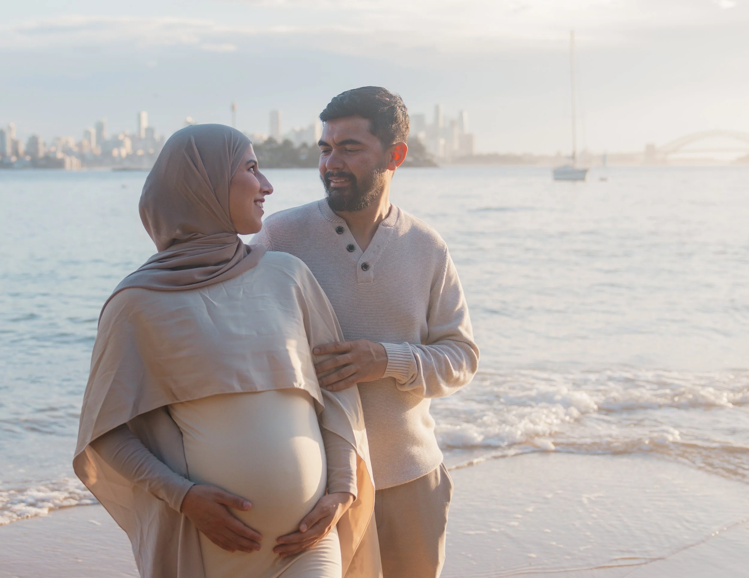 A couple on the beach, with the woman visibly pregnant, looking at each other, during sunset with city skyline and sailboats in the background.