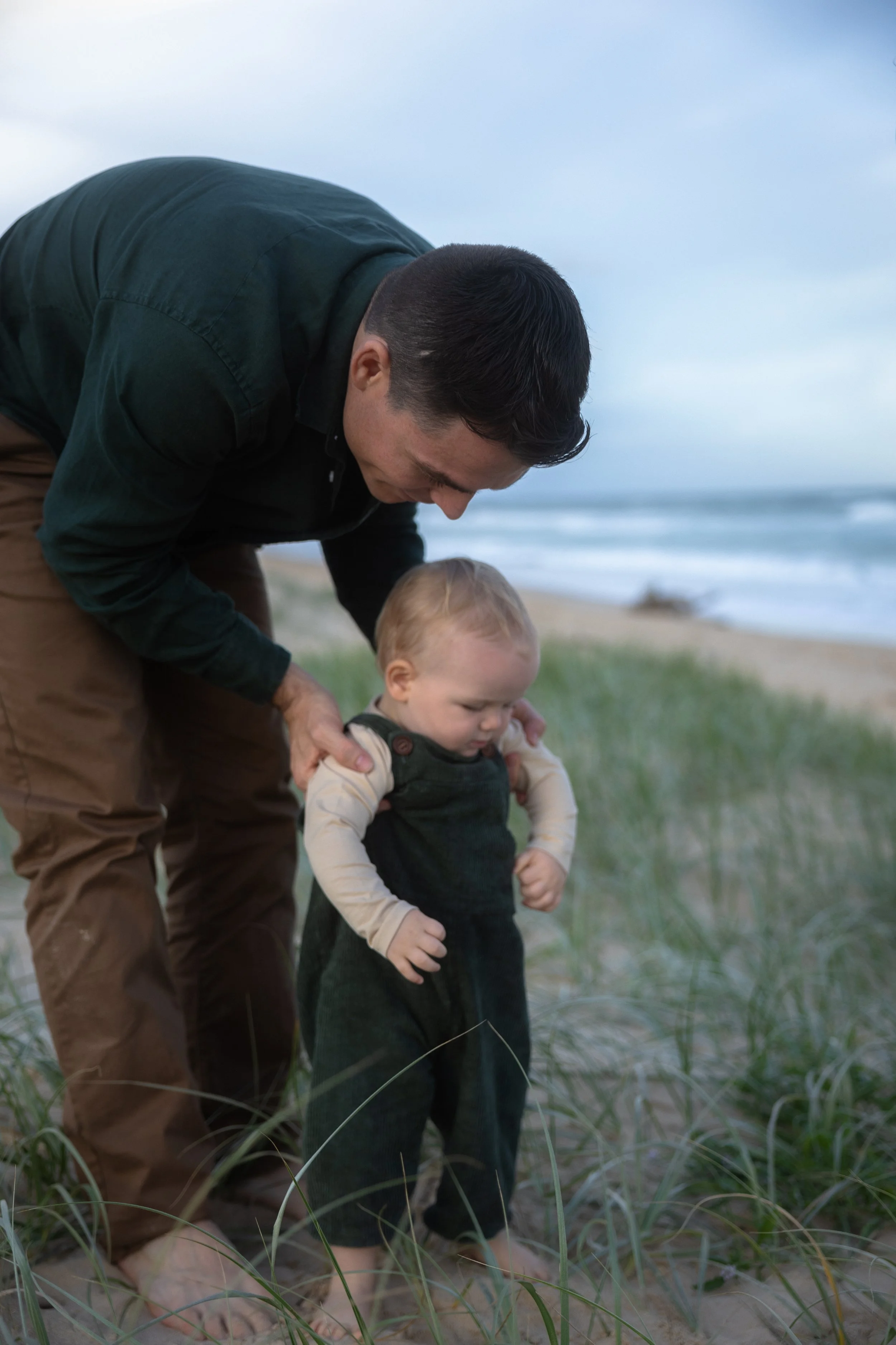 A man helping a young child walk on a sandy beach with grass, near the ocean, during cloudy weather.