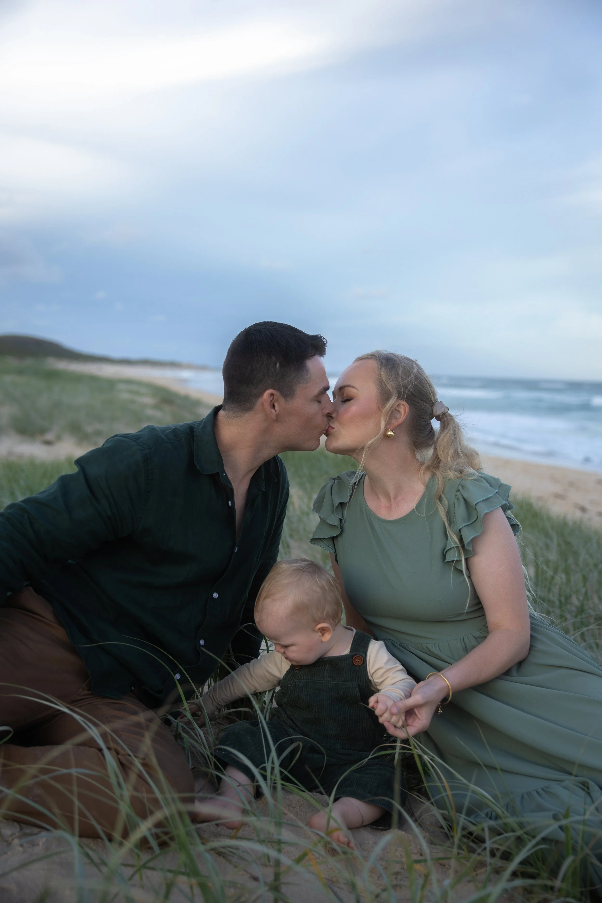 A couple sharing a kiss on a beach with a child sitting among grass and sand, ocean waves in the background, under a cloudy sky.