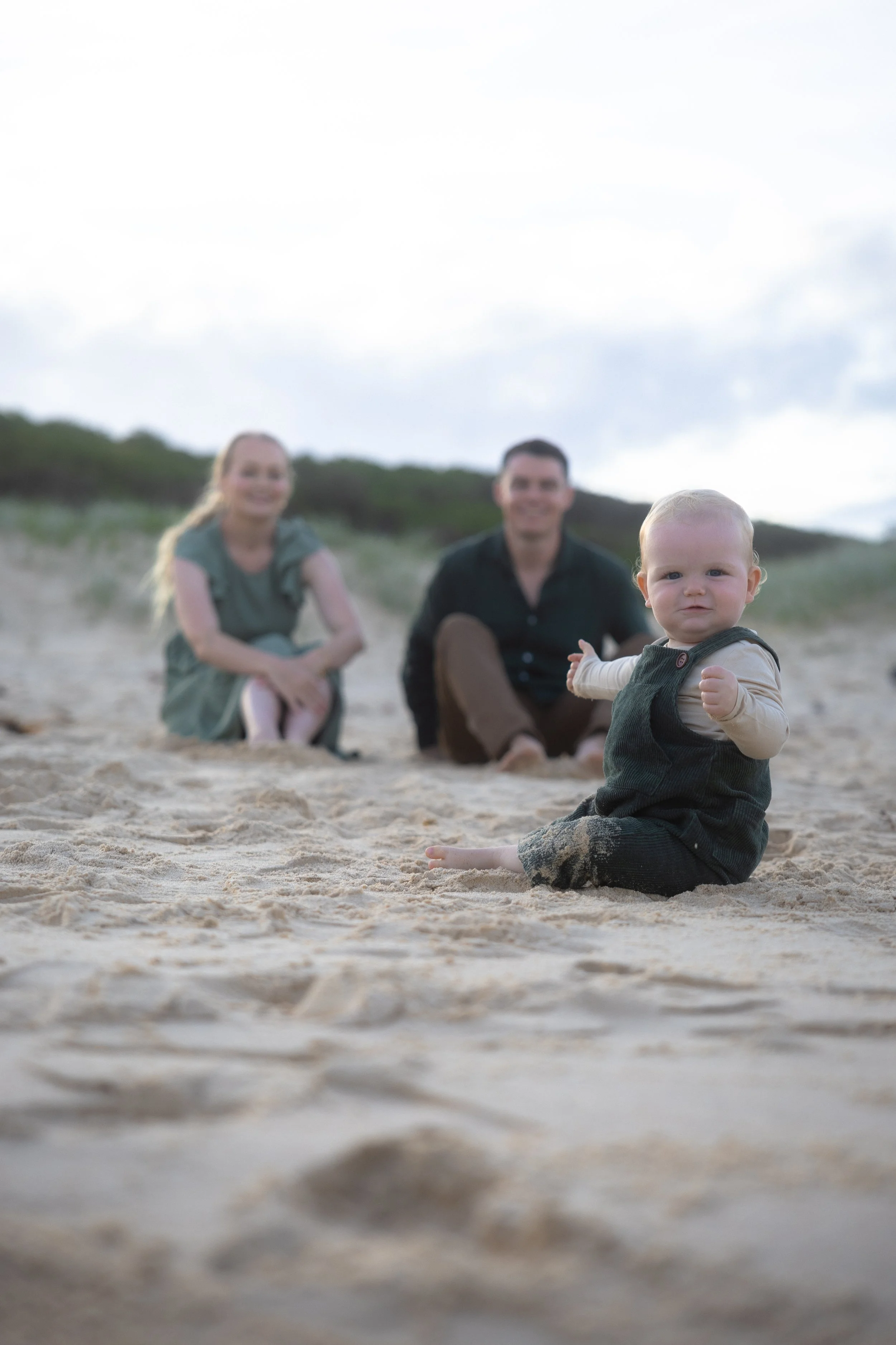 A young child sitting on the sand at the beach with sand on their clothes, with a woman and man in the background, slightly out of focus.