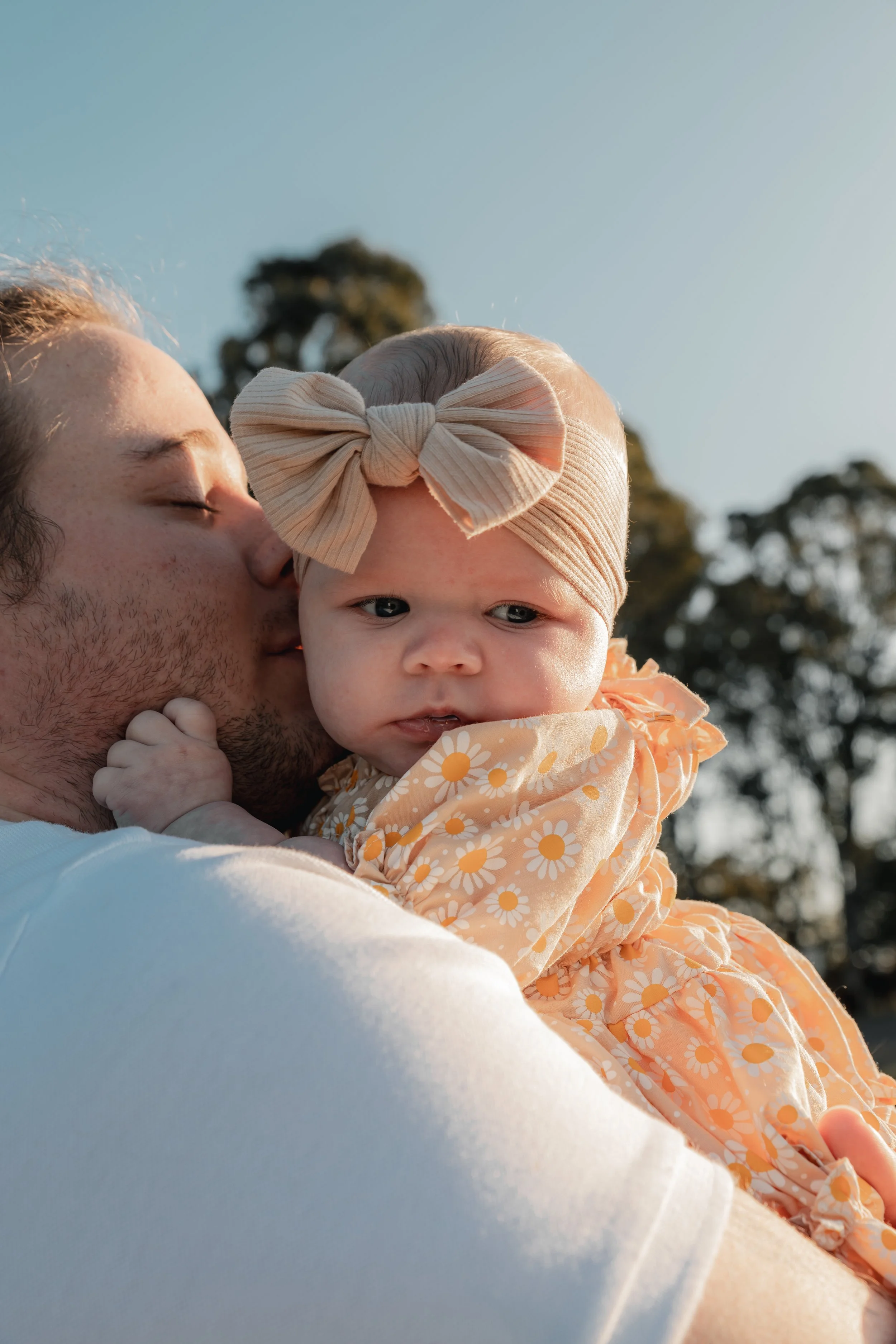 A man is holding a baby girl outdoors at sunset. The man is kissing the baby's cheek while the baby looks slightly away. The baby is wearing a headband with a large bow and a dress with a daisy pattern. The background features trees and a clear sky.